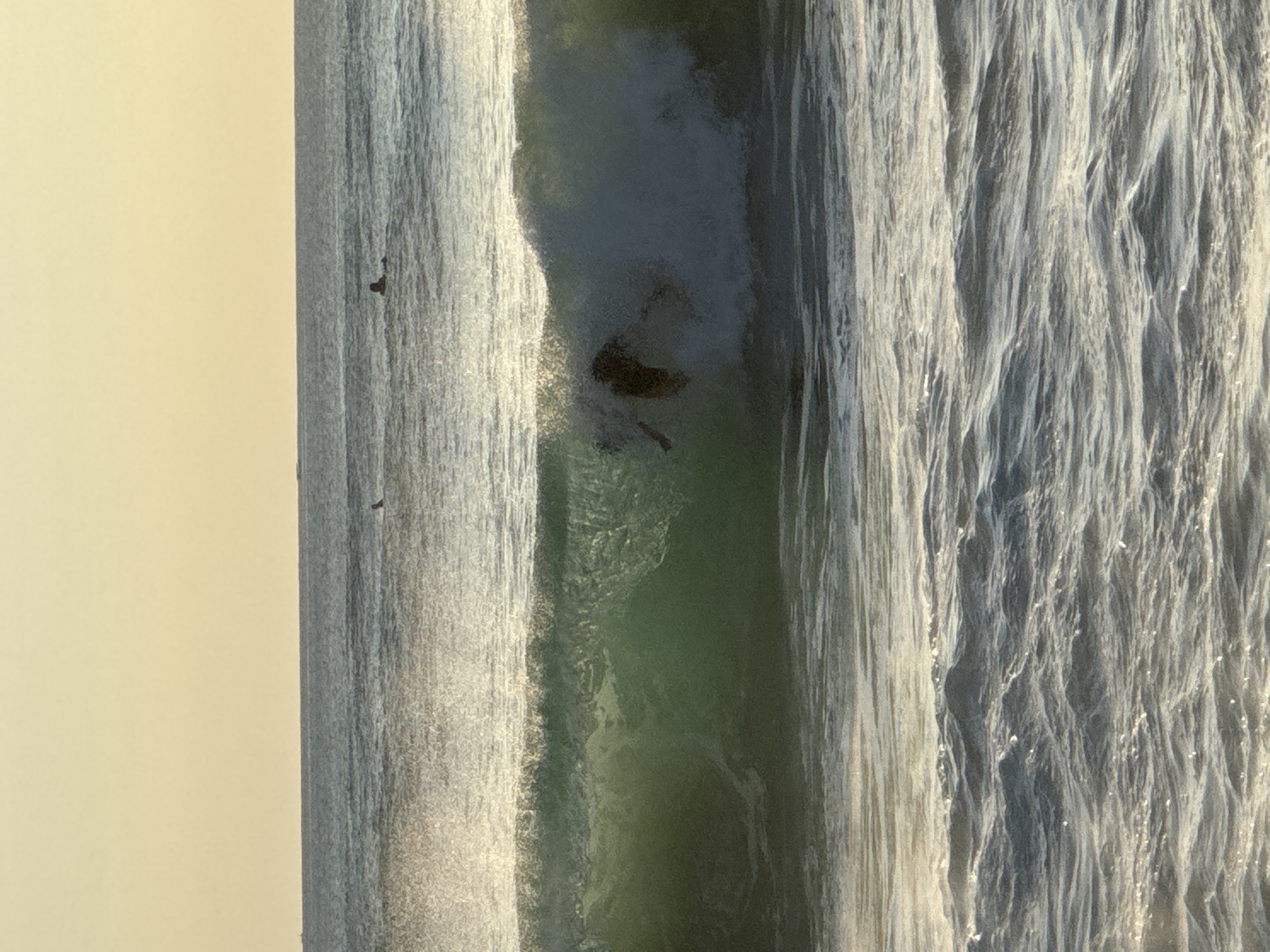 The image shows an artificial waterfall structure in Kuta Selatan, Indonesia. The artificial waterfall is composed of two vertical waterfall areas and a pool between them. Some debris is at the bottom of the pool. The backdrop is a tan-colored plain wall or sky.