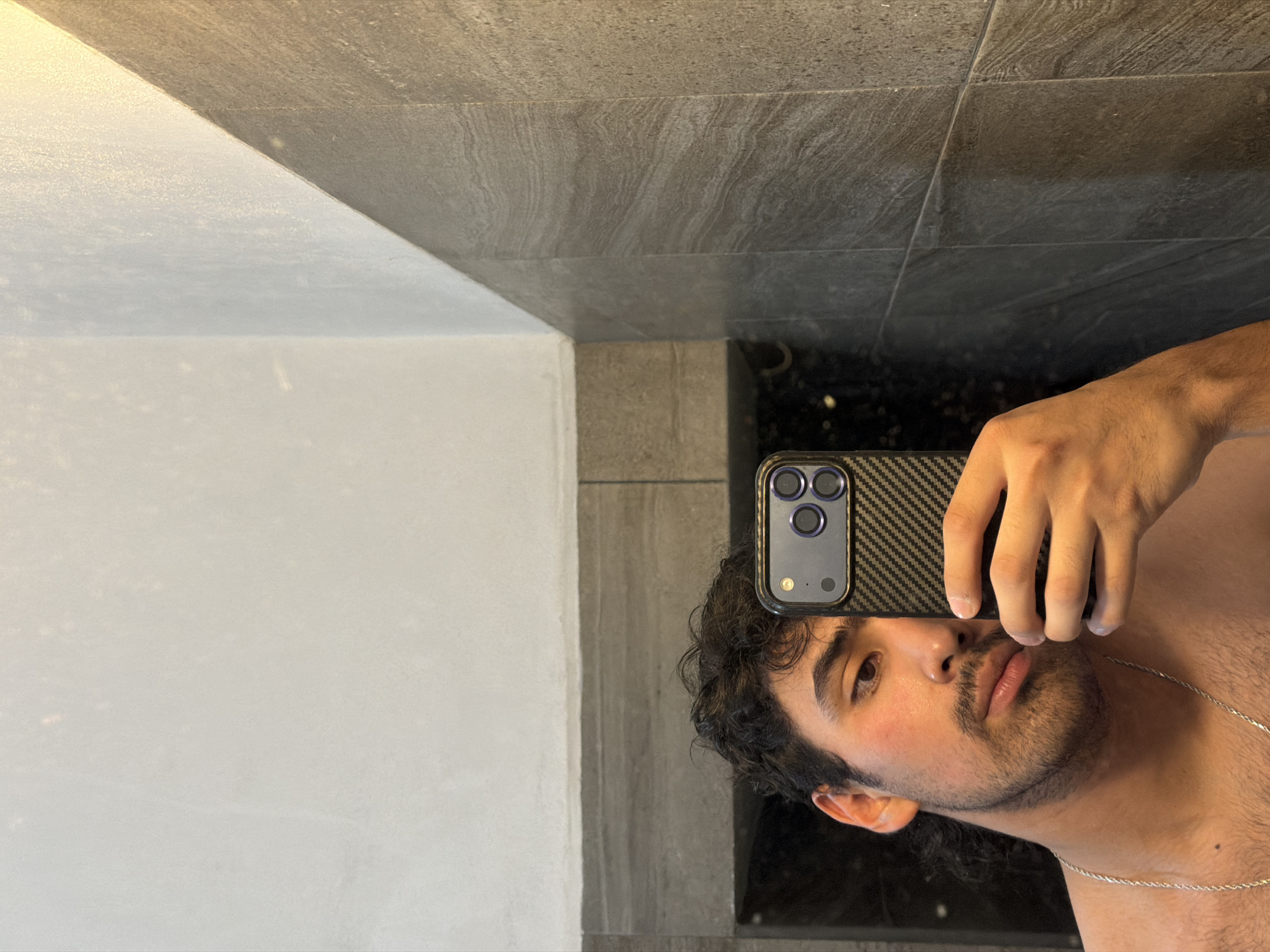 A man is taking a selfie in a bathroom. The bathroom has white and gray tile walls. The man is holding a phone with a black carbon fiber case. The man has dark curly hair and a mustache. He is wearing a silver chain. The photo's location in the city of Cuautla, Mexico cannot be ascertained from the given information.