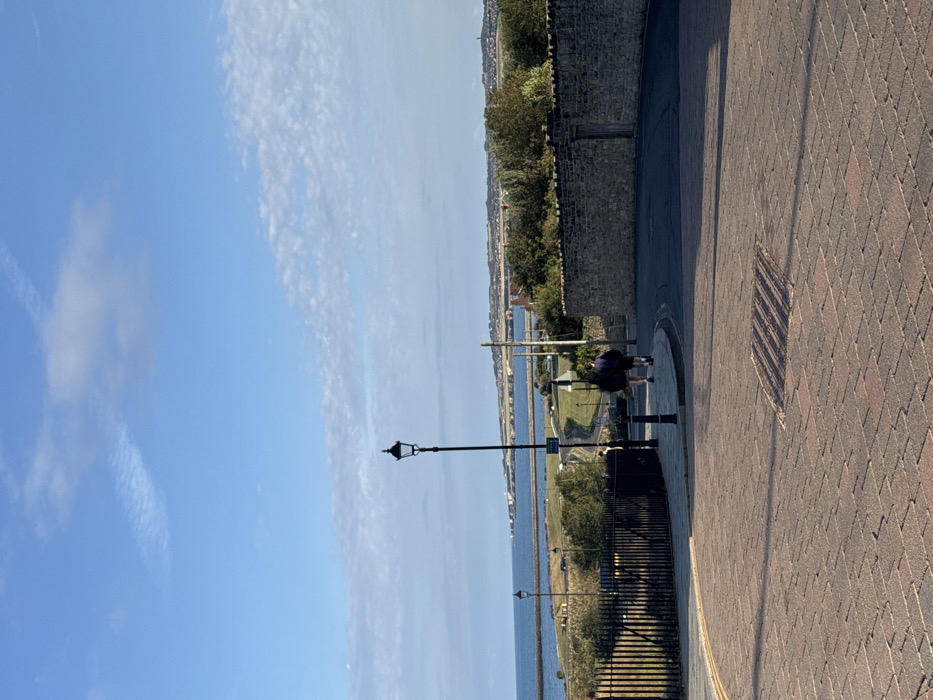 The image depicts a coastal street scene in Tynemouth, North Tyneside, United Kingdom, under clear blue skies with scattered white clouds. The sun illuminates the scene from the left, casting shadows.
In the foreground, a reddish-brown block-paved street curves to the right. A square metal grate is embedded in the pavement near the center. On the left, a black, decorative streetlamp stands on a pole, casting a shadow to its right. To the left of the streetlamp, a black metal fence or railing runs along the edge of an unseen drop. Two individuals, dressed in dark clothing, are walking away from the viewer on the right side of the street, heading around the curve.
The midground features a low, dry-stone wall on the right, which encloses a vegetated area with trees and bushes. A small, weathered wooden door is visible within this stone wall. Below the level of the foreground street, a paved path or road winds down towards the coast, with a smaller black streetlamp and a green road sign visible alongside it.
The background presents a wide expanse of calm blue sea, extending to the horizon. A breakwater or pier structure is visible projecting into the water on the left. Beyond the breakwater, a distant coastline with prominent cliffs and white structures, possibly a lighthouse (Souter Lighthouse), is visible. Further along the horizon, a town or city with numerous buildings and green hills is present.