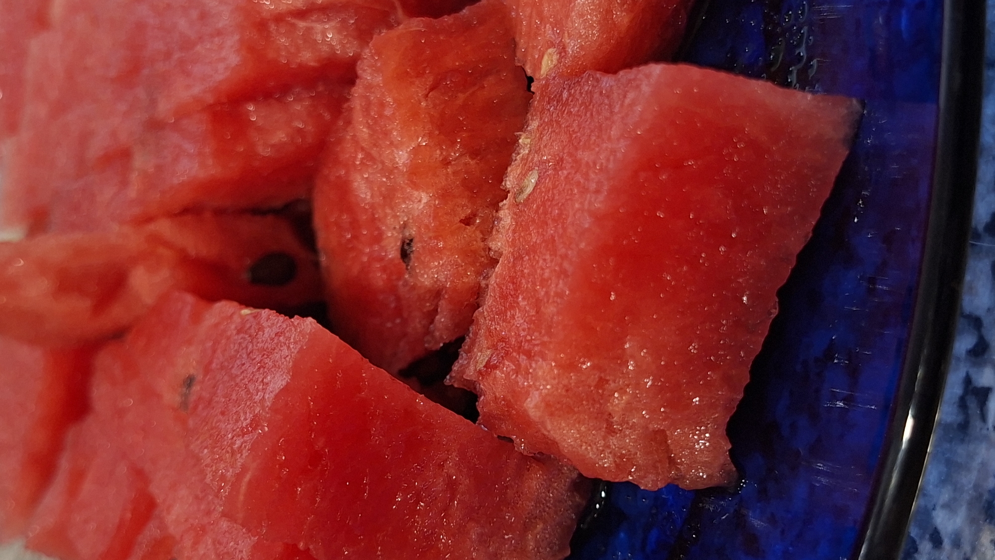 The image is a close-up, overhead view of several cubed pieces of red watermelon resting on a dark blue plate. The watermelon pieces vary in size and shape, with some showing visible seeds. The plate has a glossy surface, and there are a few droplets of water or juice visible on it. The background is blurred, focusing the viewer's attention entirely on the fruit. There are no people or identifiable landmarks visible in the image. The context of Myadzyel, Belarus is not supported by any visual information in the provided image.