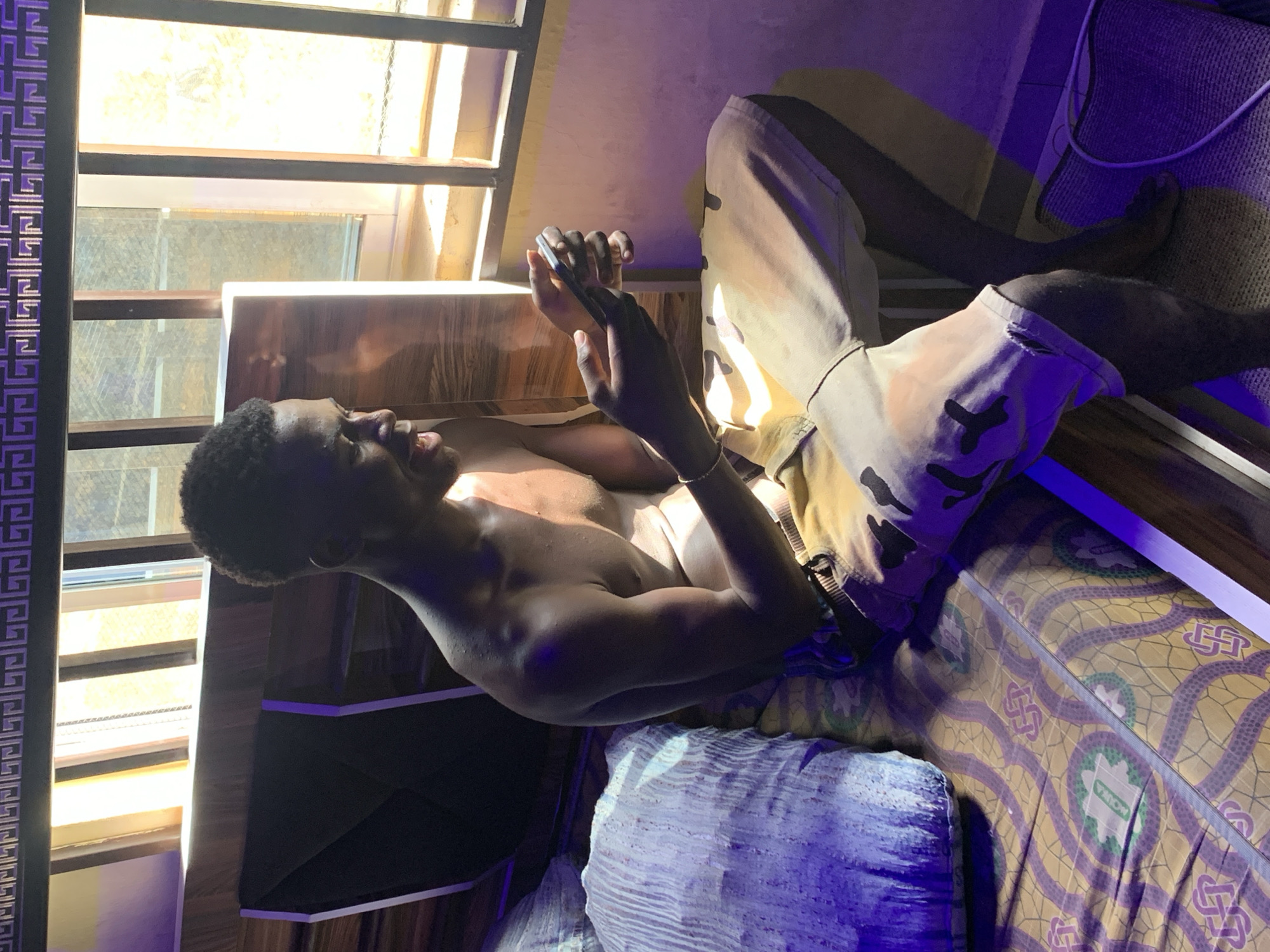 A young, dark-skinned man is reclined on a bed, seemingly in an indoor setting. He is shirtless and wearing camouflage pants. He is looking at and holding a mobile phone with both hands. The bed has a patterned bedspread and a blue pillow. A window with bars is visible, and light streams in. There is what appears to be a decorative panel with a repeating pattern on the left side of the frame. A bed is pushed against the wall next to a window. The cables of a device hang behind the man, and the objects give the impression that the photo was taken indoors. The specific location within the city of Lokoja, Nigeria, is impossible to pinpoint without further information.