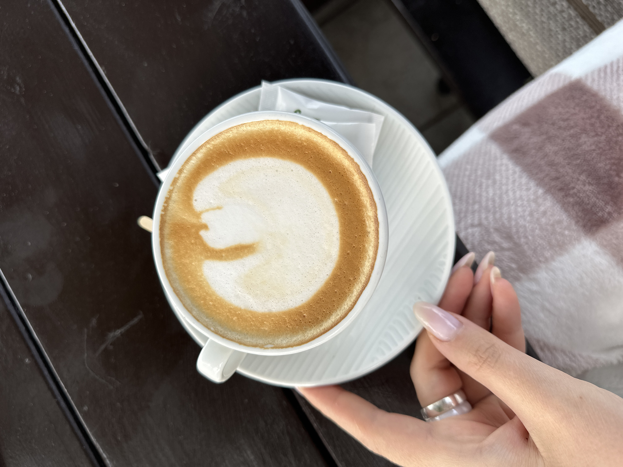 A cappuccino sits in a white cup on a matching saucer atop a dark brown wooden table. A hand, adorned with a silver ring and long nails, rests near the saucer. A beige and white plaid blanket is partially visible in the background. The location is inferred to be Šušanj, Montenegro, based on the implied setting.