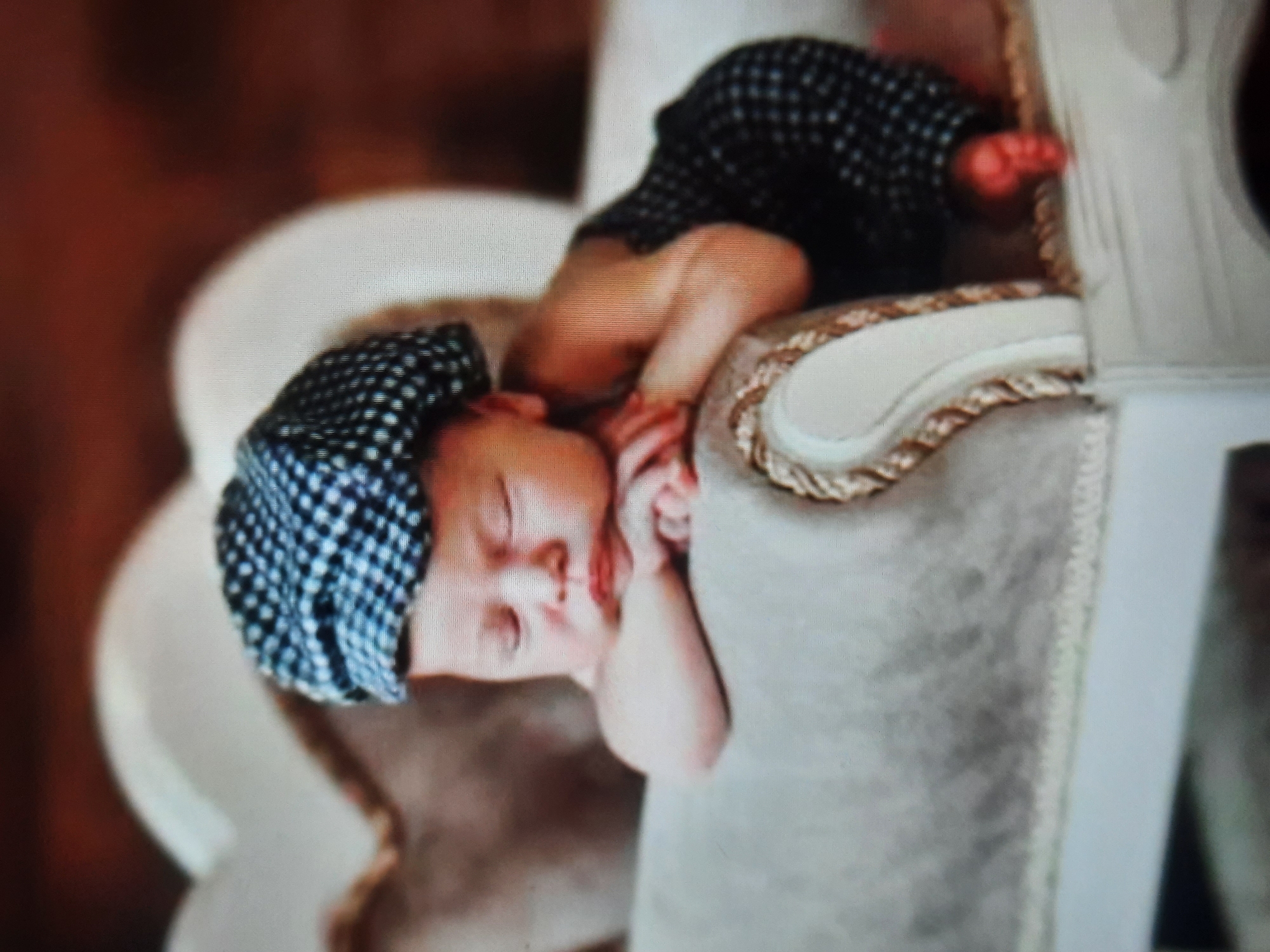 A newborn baby is sleeping on a plush couch, wearing a black and white checkered cap and matching overalls. The baby's head is resting on its folded arms, with eyes closed and mouth slightly open. The background is blurred, showing warm, brown tones and a light-colored upholstered headboard or cushion. The couch has a decorative trim along the armrest. The image appears to be a professionally taken photograph, likely in a studio setting or a well-decorated home. There is no information provided that links this image to the city of Rosenheim, Germany.