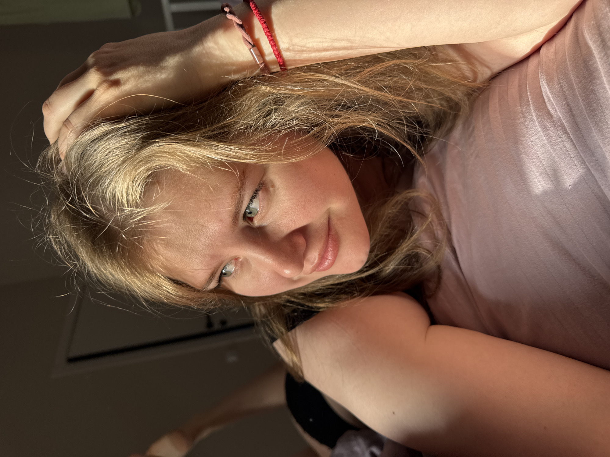 The image captures a close-up portrait of a young person, likely female, with long, wavy blonde and light brown hair, lying down indoors. She looks directly at the camera with a gentle, relaxed smile and light-colored eyes. Her left arm is bent, with her hand positioned near her head, and she wears two hair ties or bracelets on her wrist: one red string and one lighter pink/brown.
She is dressed in a light pink, subtly ribbed top, with a hint of a dark garment visible beneath. The scene is illuminated by strong, directional natural light, likely sunlight streaming in from a window, which creates bright highlights on her hair, skin, and clothing, and casts soft shadows, suggesting either early morning or late afternoon. The background is dark and indistinct, indicating an indoor setting, possibly a room. The overall mood conveyed is calm and inviting. No text is visible in the image. The location context provided for this image is Ферма, Russia.