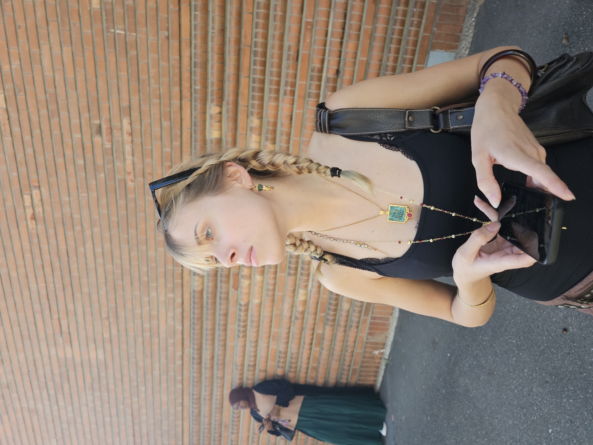 The image depicts a woman with blonde hair in braids, wearing a black tank top and multiple necklaces, holding a smartphone. She is standing in front of a brick wall. In the lower portion of the image, there's another person visible, wearing a head covering and a long skirt. The location appears to be an urban setting. The information available does not indicate the specific location within the city of Massy, France.