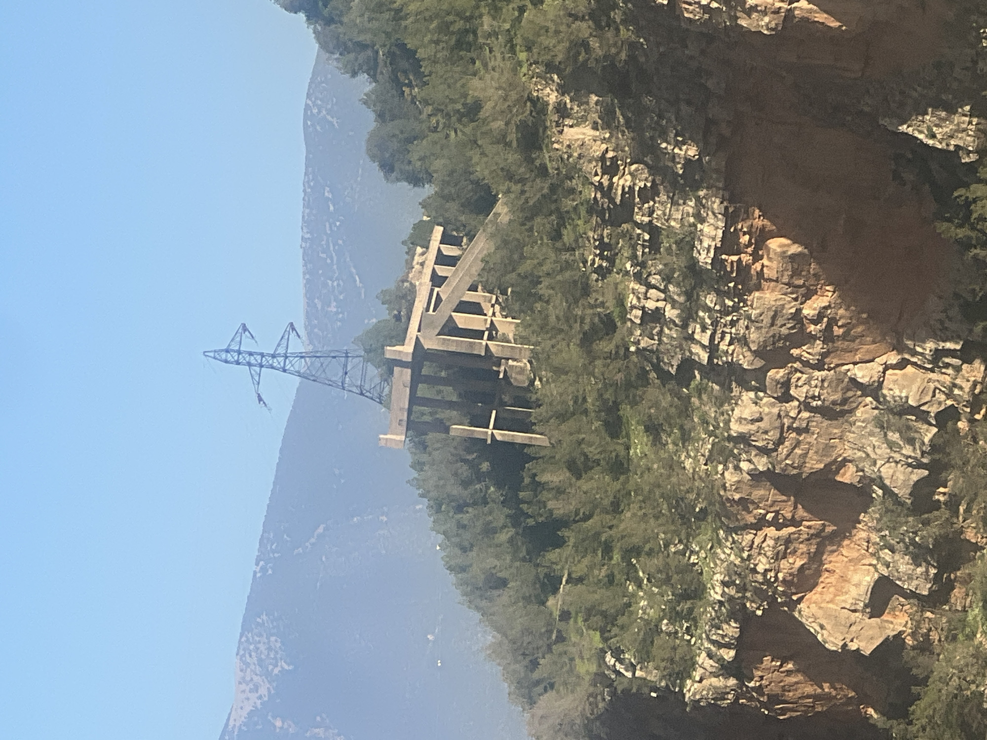 The image shows a steep, rocky hillside with vegetation near Bin El Ouidane, Morocco. A concrete structure with a power line tower stands on the hillside. Behind the structure and tower, there is a mountain range under a clear blue sky. The foreground shows more of the rocky terrain.