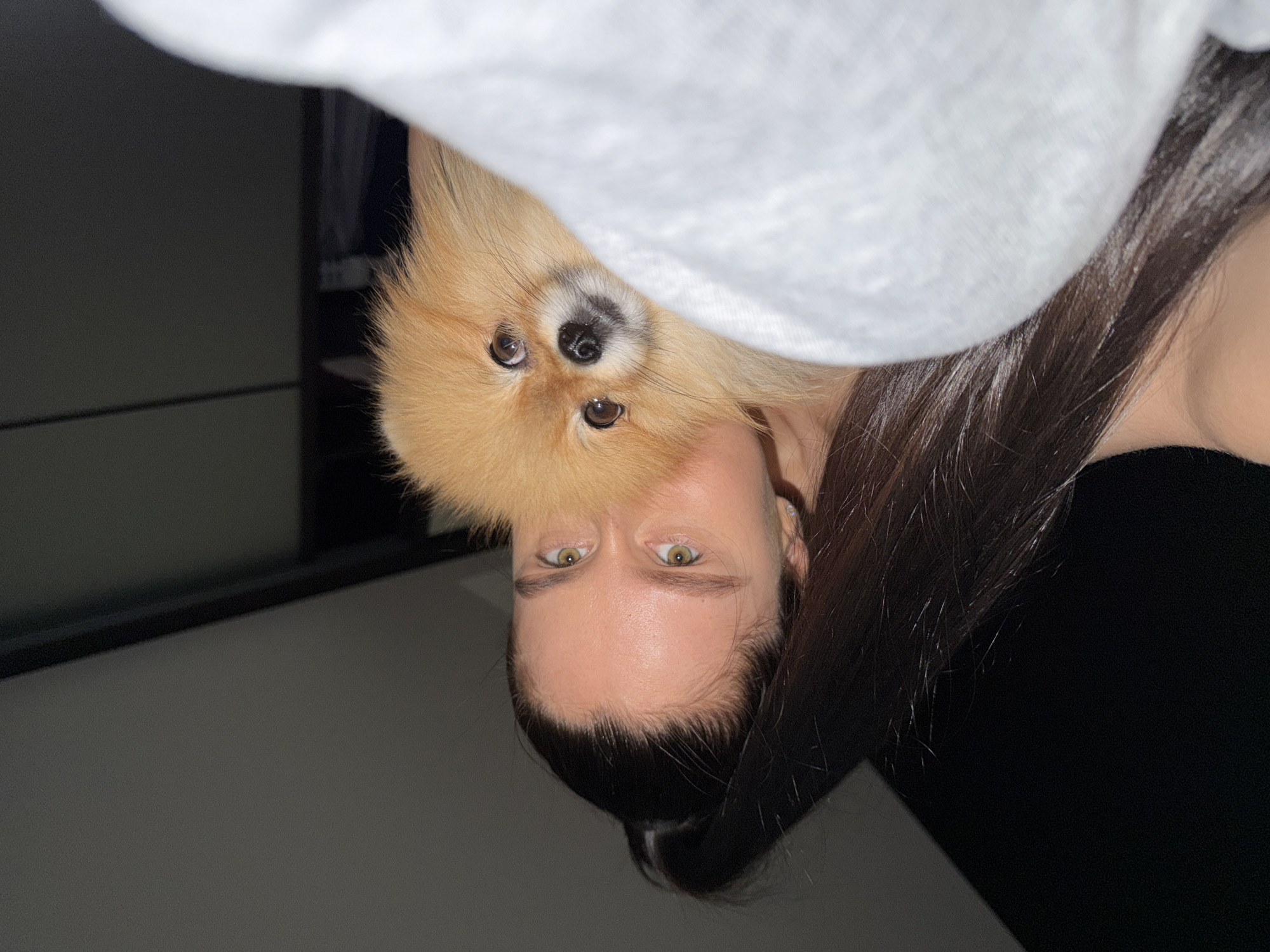 A selfie depicts a woman with dark hair pulled back and a tan Pomeranian dog. The woman has light skin and green eyes. A light gray fabric drapes over the top portion of the frame. The background includes a black cabinet or furniture piece and dark surroundings. The location is not discernible from the image, so I cannot confirm it to be Khimki, Russia.