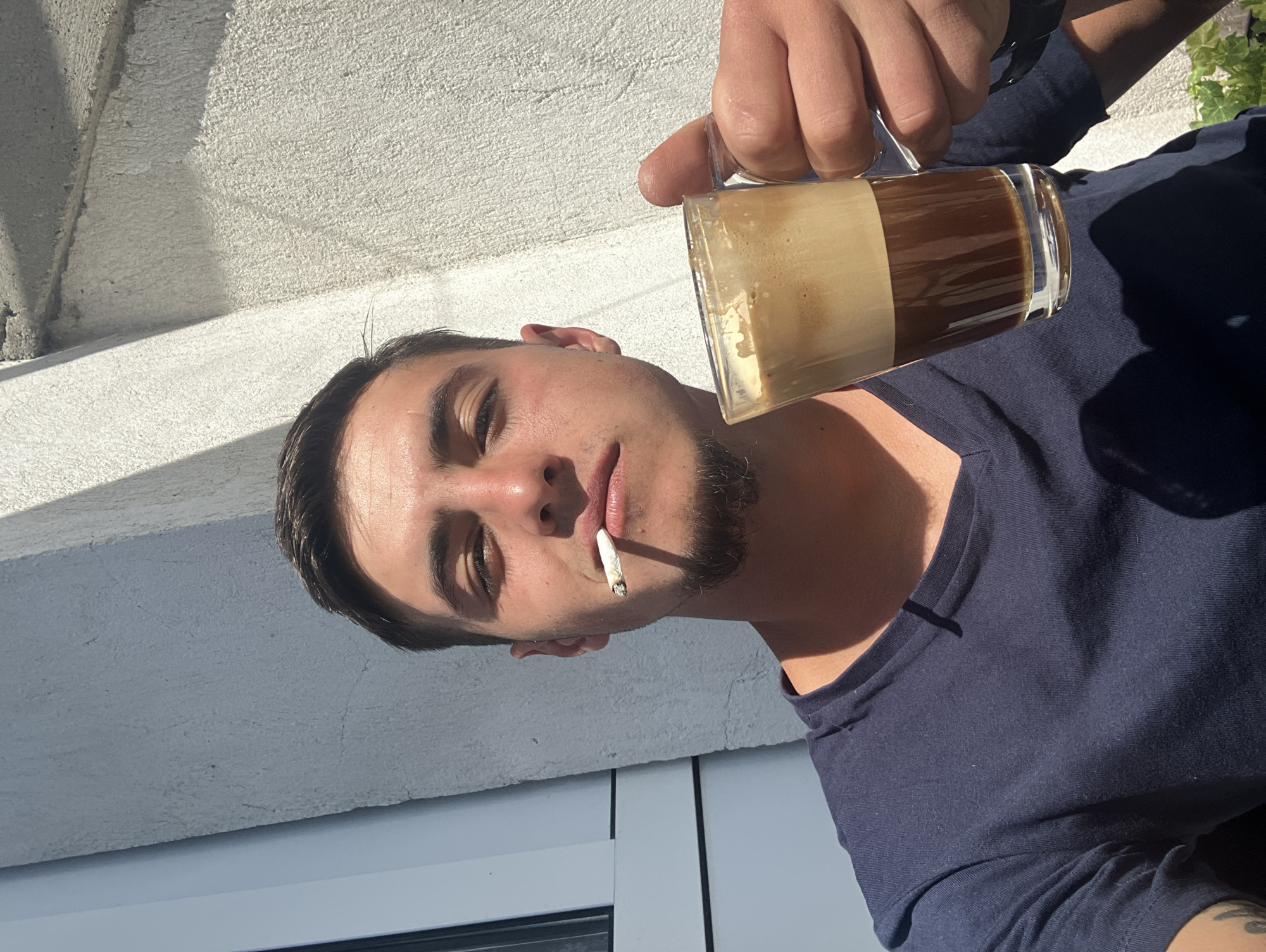 A man with short, dark hair and a trimmed beard holds a glass mug filled with a two-layered coffee drink (likely a latte or cappuccino) in his right hand. A cigarette is in his mouth. He is wearing a dark blue t-shirt and a watch on his left wrist. The background includes a white exterior wall with horizontal seams. The location is Krastevich, Bulgaria.