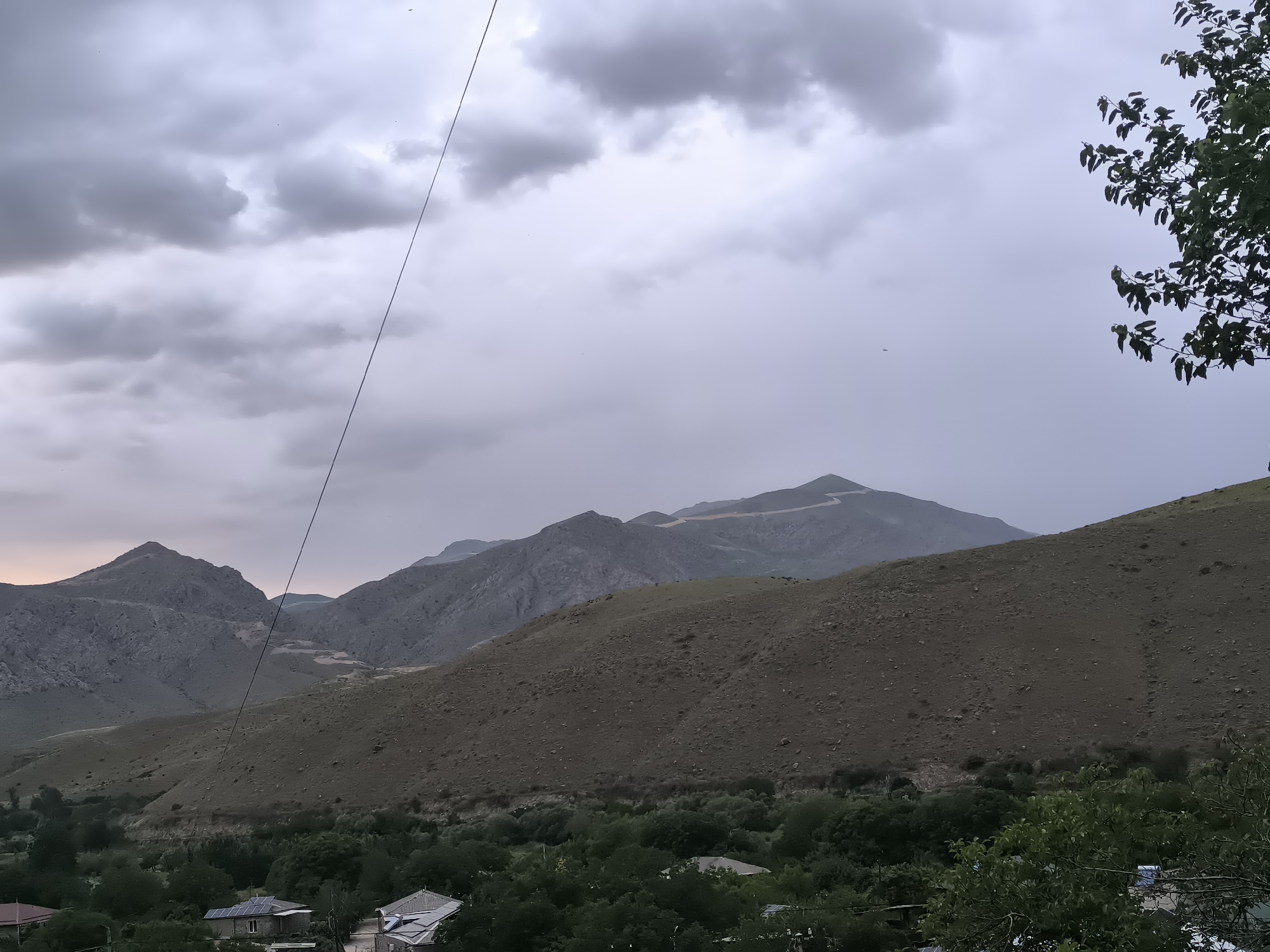 This image captures a panoramic view of a mountainous, rural landscape in Areni, Armenia, under an overcast sky. The scene is dominated by a series of rugged mountains in the background, their peaks and slopes appearing rocky and sparsely vegetated. A winding path or road is discernible on one of the higher mountain ridges.
In the mid-ground, rolling hills, covered in dry, brownish-green grass and scattered rocks, descend towards a valley. The valley floor is densely forested with lush green trees, which partially obscure a few buildings nestled within. One notable detail is a building visible in the lower left, featuring several solar panels on its roof, suggesting a reliance on renewable energy. Another building has a distinctive corrugated metal roof.
The sky is heavily overcast with thick, grey clouds, creating a subdued and diffused light across the landscape. The weather appears to be either pre-storm or just after rainfall. A dark line, likely a power line or cable, cuts diagonally across the upper portion of the image. In the upper right, the dark green leaves of a tree branch partially frame the scene. Small, indistinct specks, possibly birds or insects, are scattered in the sky. No human activity or visible text is present in the image. The time of day appears to be either late afternoon or early evening, given the soft, muted light.