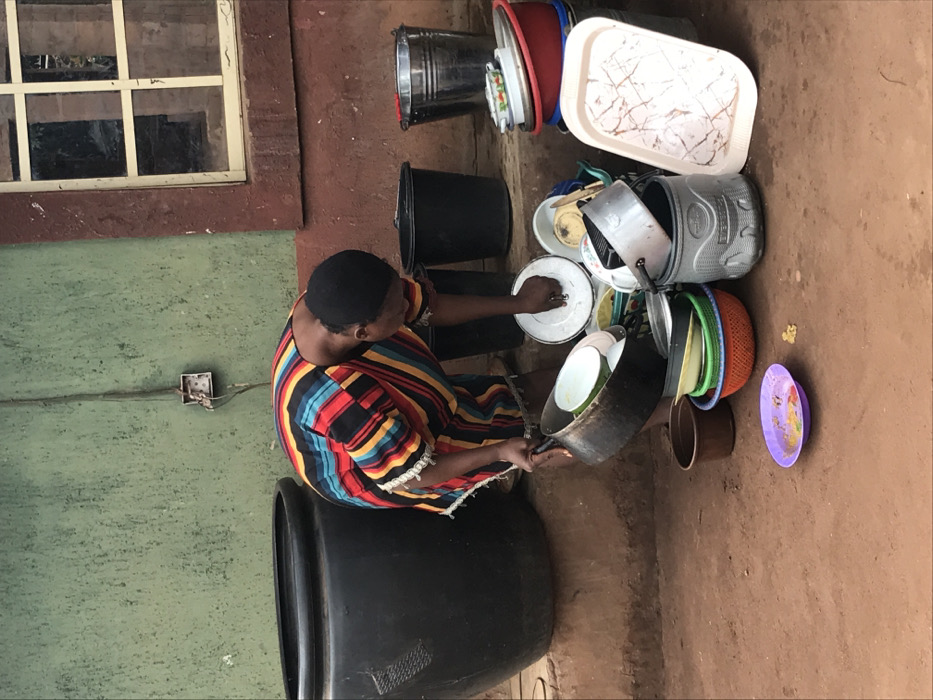 A woman in a colorful striped dress washes dishes outdoors. She sits on a large black container while cleaning pots and plates. A stack of various kitchenware including buckets, bowls, and trays is piled nearby. The scene is set against a backdrop of a weathered wall with a window and exposed electrical outlet. The ground is a reddish-brown dirt.
