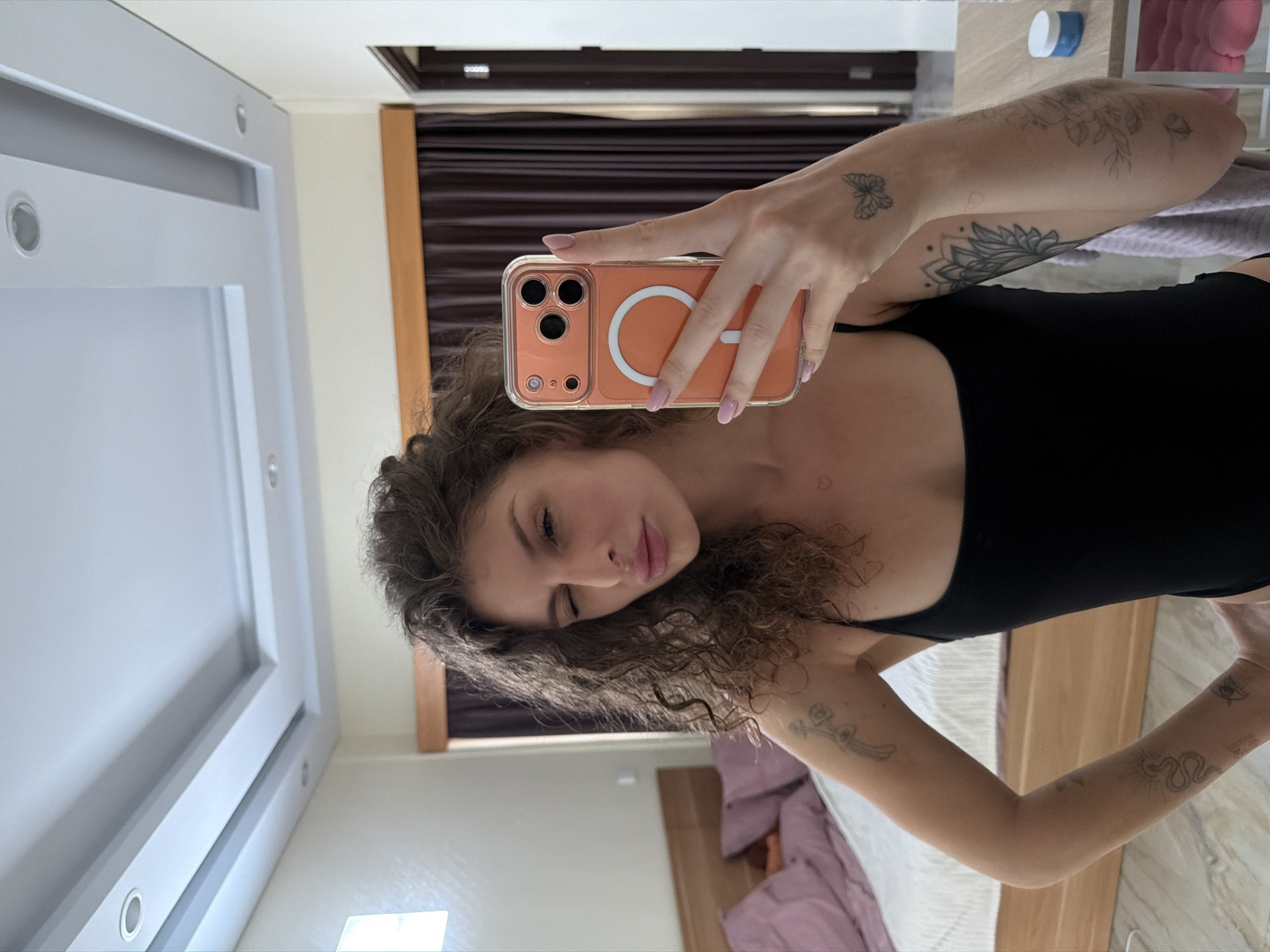 A young adult female with curly brown hair is taking a mirror selfie in what appears to be a bedroom. She is wearing a black tank top and has several tattoos visible on her arms, including a butterfly, a flower, and a snake. Her right hand is holding an orange phone with a clear case. She is winking at the camera. The room has a bed with purple pillows, a wooden headboard, and a modern ceiling with recessed lighting. Dark brown curtains are visible in the background, and a wooden door is partially seen on the right. A table with a white jar is also present. The floor is tiled. The lighting suggests it is daytime. This scene is located in North Kuta, Indonesia.