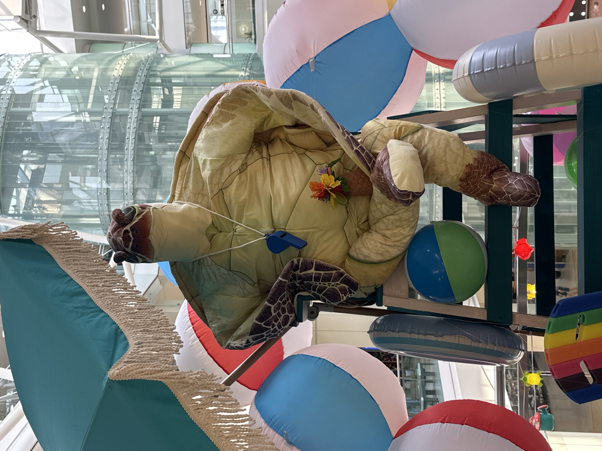 This is a medium shot, eye-level, indoor photograph of a large, stuffed turtle posed on a green step ladder amidst a collection of inflatable beach balls and other summer-themed props. The turtle is wearing a yellow costume that resembles its shell and skin. A blue whistle on a white cord hangs around its neck. A small vase with artificial flowers is tucked into its arm. To the left, a teal umbrella with fringed trim is visible. Surrounding the turtle are various inflatable objects: large, multi-colored beach balls, a gray and white striped inflatable tube, and a two-toned blue and green inflatable ball. The background shows a modern, multi-story building with a cylindrical glass elevator shaft and reflective surfaces. This scene is located in the atrium of the Lougheed Town Centre in Burnaby, Canada.