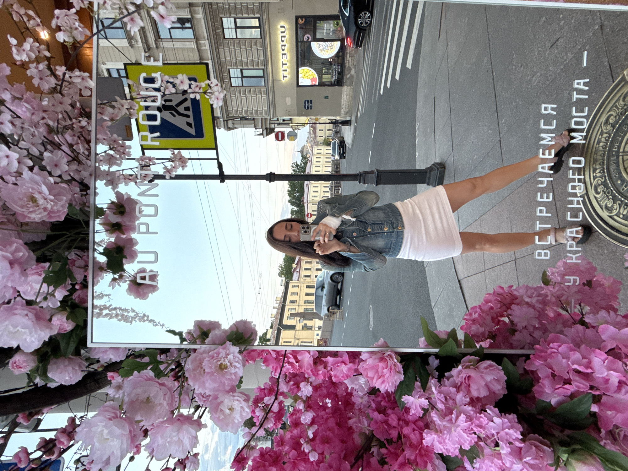 A woman is captured taking a mirror selfie on an urban street, likely in Saint Petersburg, Russia, based on the textual cues. The large rectangular mirror is framed by an abundance of pink artificial flowers, resembling peonies and cherry blossoms, which partially obscure the reflection and foreground.
The woman, with long dark hair, is wearing a denim jacket over a white top and a white skirt or dress. She holds a smartphone to take her picture. The mirror reflects a bright daytime scene, showing an old, multi-story building with numerous windows across the street. A shop sign for "TITIPUU" (reflected upside down) is visible on one of the buildings. A black car is parked on the street, and a lamppost stands in the middle ground. A yellow pedestrian crossing sign with a black symbol is also clearly reflected.
Visible text on the mirror includes "PONT ROUGE" (partially visible and possibly a stylized name, given the context, potentially linking to the "Red Bridge") and the Russian phrase "ВСТРЕЧАЕМСЯ У КРАСНОГО МОСТА," which translates to "Let's meet at the Red Bridge." To the right, an ornate round metal plate with intricate patterns is on the ground. The weather appears clear and sunny, suggesting a pleasant day. The scene combines a personal moment with the bustling urban environment, decorated with a touch of artificial nature.