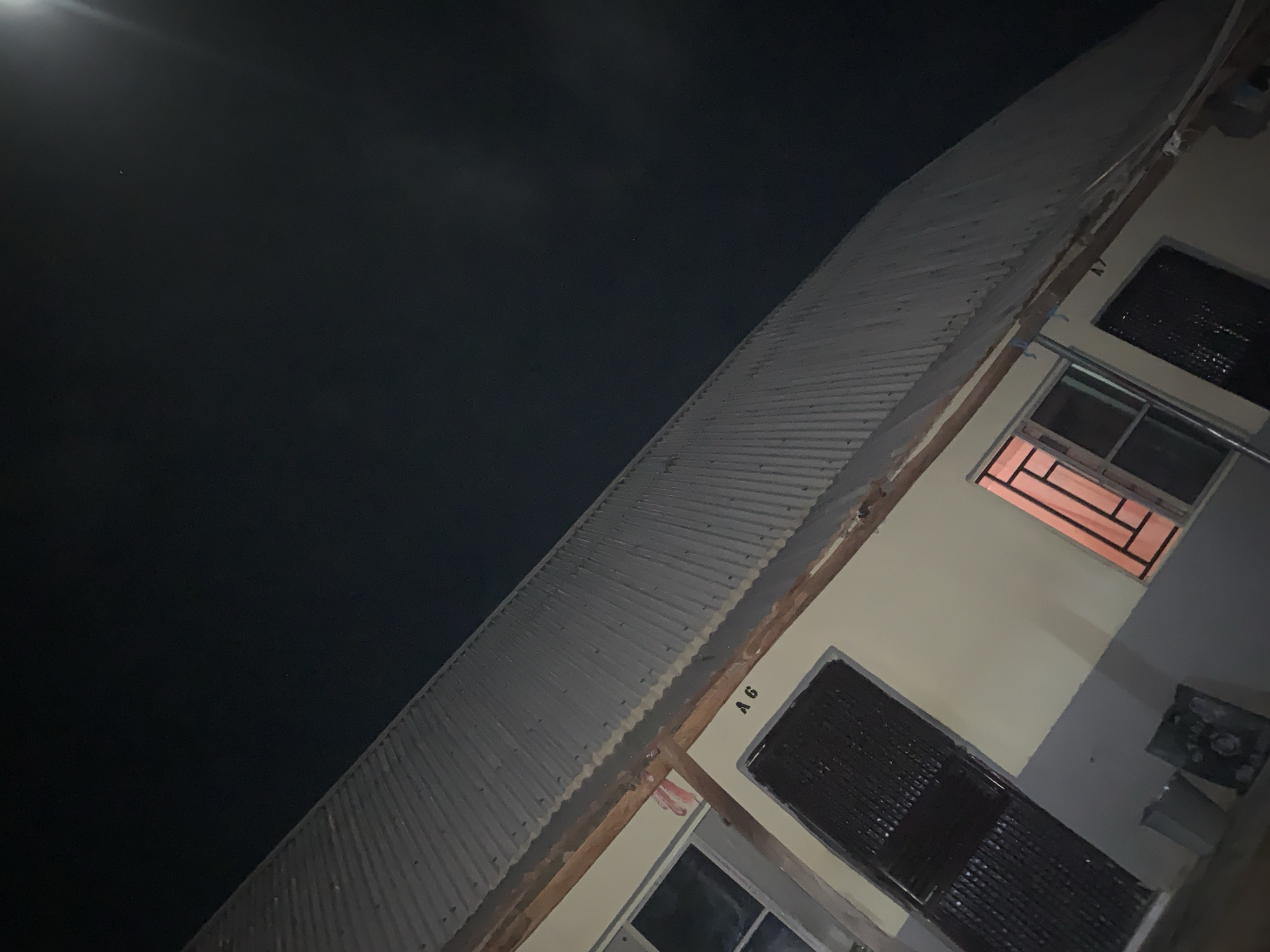 The image presents an angled view of a building at night, likely located in Keffi, Nigeria, as that information was given to me. The sky is dark and clear with a bright moon. The building has a corrugated metal roof. The exterior wall is light-colored and features multiple windows and a door. A window, located in the upper portion of the wall, emits an orange glow from inside. A label "A6" is visible to the side of one of the window. An electric meter box is visible on the wall on the bottom right.
