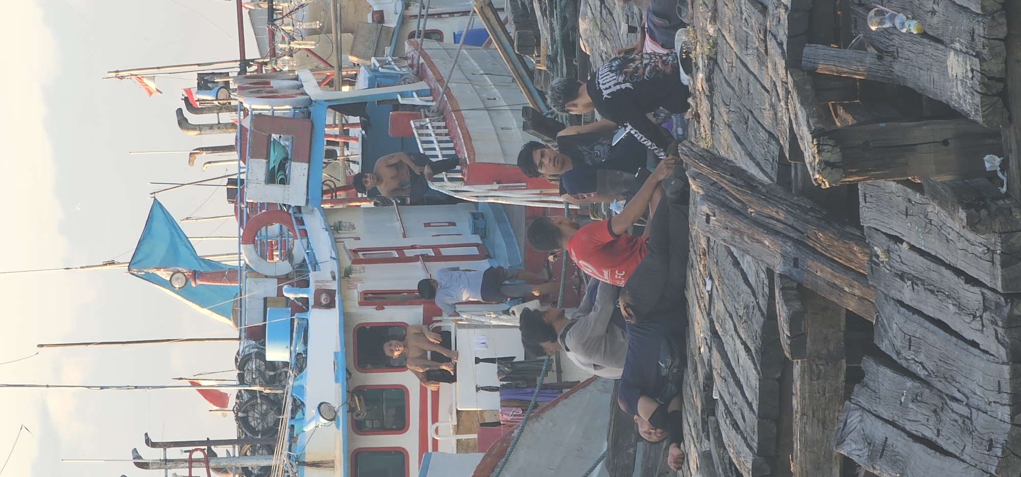 The image captures a scene at the port in Denpasar, Indonesia. Several colorful fishing boats are moored alongside a weathered wooden pier. Several people are visible; some are aboard the boats, while others are on the pier. Some of the men on the boats are shirtless. The pier is made of dark, aged wooden planks.