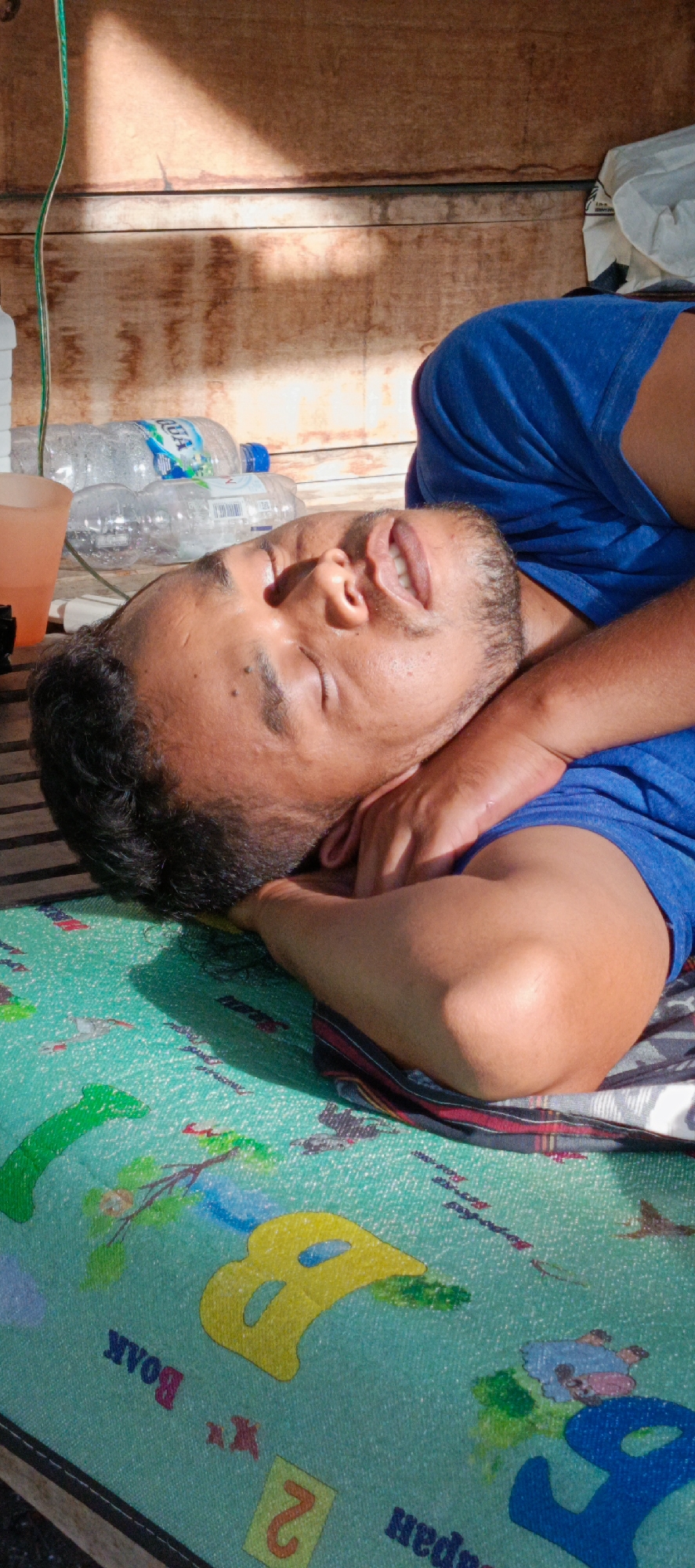 A man is sleeping on his side, on a colorful mat, outdoors. He is wearing a blue t-shirt and his head is resting on his arms. His eyes are closed and his mouth is slightly open. In the background, there are wooden planks and some plastic water bottles. A shadow from above is cast across the wooden planks. The mat he is sleeping on has various colorful shapes and letters on it, including what appears to be the letter "B". The overall scene suggests a casual, relaxed setting, possibly a park or a backyard, in a location that would be warm enough to sleep outdoors. This image was taken in Pashiebeutong, Indonesia, a place known for its tropical climate.