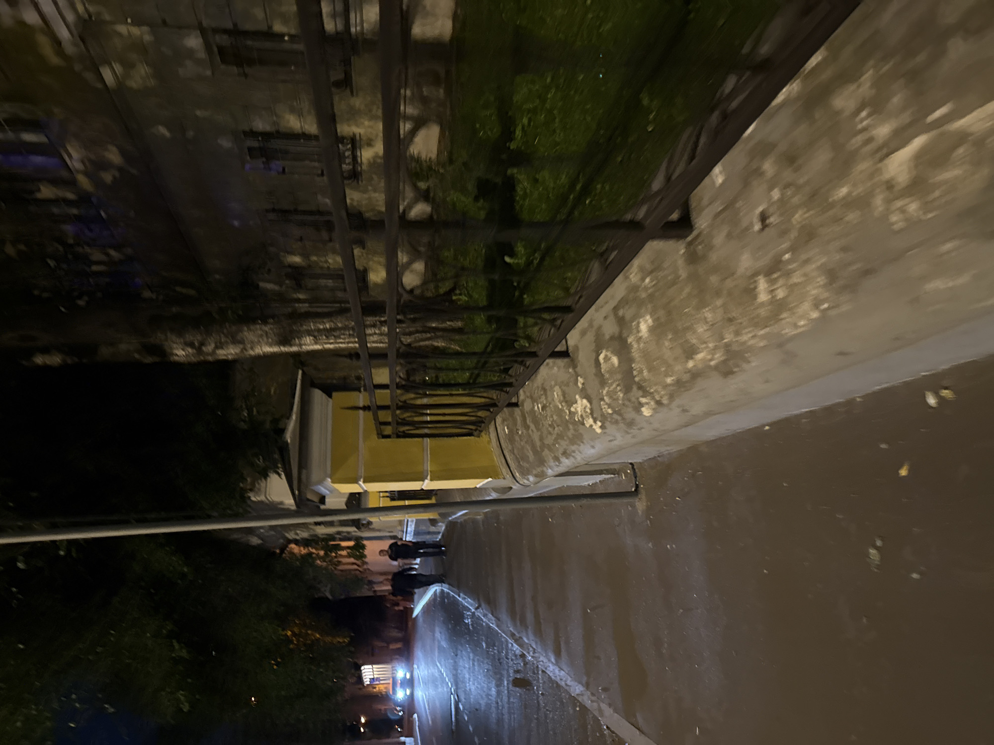 The image presents a nighttime street scene in Moscow, Russia, observed from a low, pedestrian-level perspective looking forward along a sidewalk. In the immediate foreground on the right, a textured, weathered concrete wall extends horizontally, topped by a black wrought-iron fence featuring decorative circular and curved motifs. Behind the fence, a narrow strip of dark green grass is visible. Further back, a two-story building with bright yellow walls and white architectural trim, including a prominent corner element with a capped roof, stands. A grey utility pole is positioned near the corner of this building.
On the left, a wide, dark asphalt sidewalk extends into the distance, appearing damp or reflective under the ambient lighting. Two male individuals, both dressed in dark clothing, walk away from the camera on this sidewalk. Parallel to the sidewalk, a wet asphalt road is present. A white vehicle with illuminated headlights is visible approaching in the far distance, casting bright reflections on the road surface.
The background is dominated by dark, dense tree foliage on both sides of the street, partially obscuring the upper portions of buildings. Behind the yellow building and trees, a larger, darker multi-story building with numerous windows is dimly discernable. The scene is illuminated by various artificial light sources, primarily from streetlights and the vehicle's headlights, which create areas of strong reflection and deep shadows.