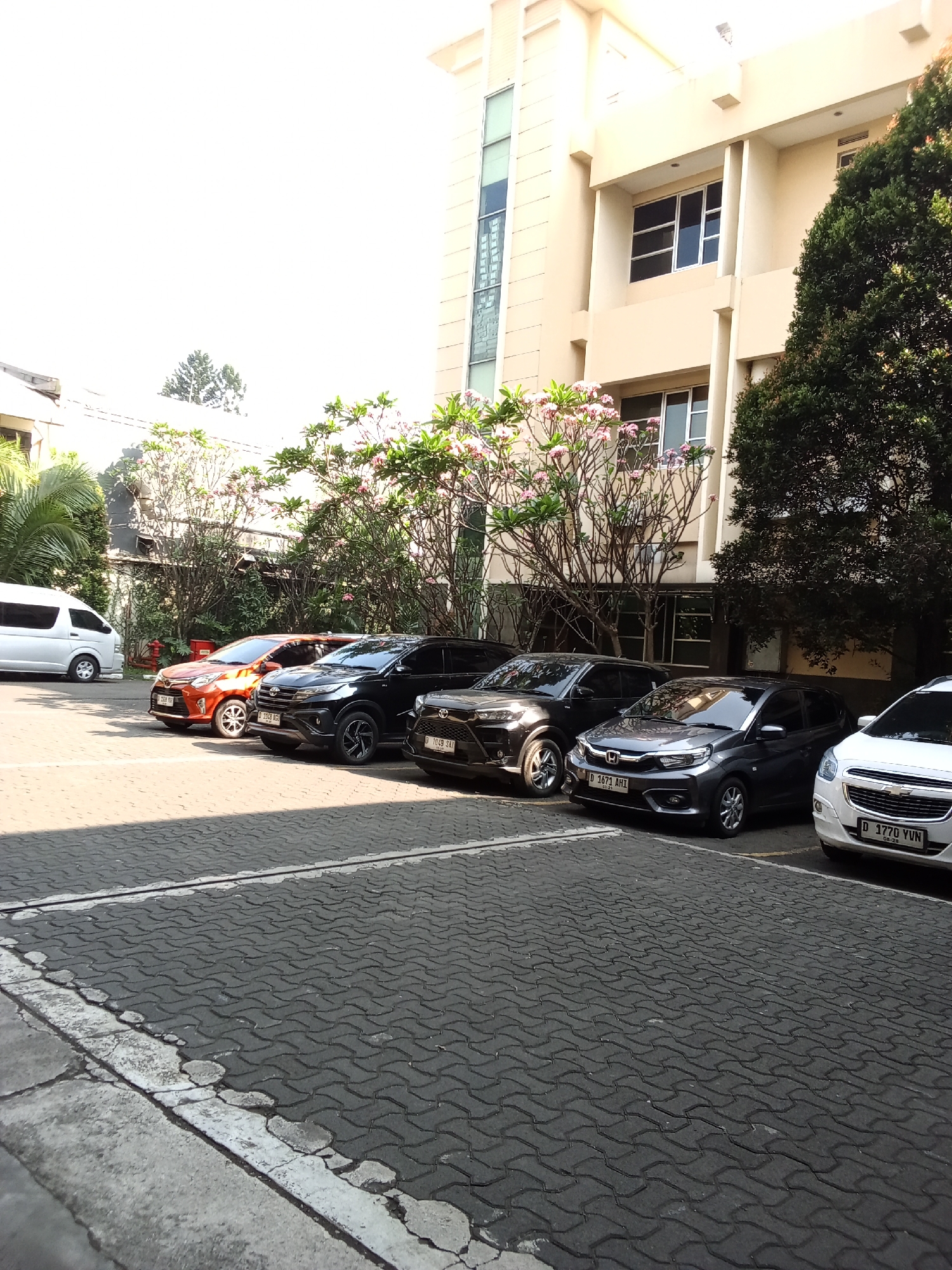 A parking lot is situated in front of a tan, multi-story building. The building has several windows and is partially obscured by lush green trees and flowering pink bushes. In the foreground, there is a paved area with interlocking gray bricks, bordered by concrete and white painted lines. Parked cars are visible on the paved surface. A white van is parked to the left. Several cars are lined up from left to right: an orange sedan, a black SUV, a dark gray hatchback, and a white sedan at the far right. The sky above is bright and appears to be clear.