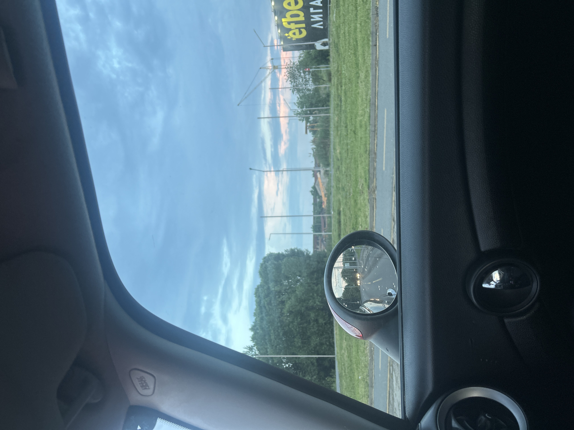 The image shows a view from inside a car, looking out the passenger side window. The exterior environment consists of a road, a grassy area, and a large billboard with the word "efbet" visible on it, written in Cyrillic script. The sky is visible above, displaying a cloudy sky at dusk with hints of pink. The side mirror of the car reflects the road behind, showing other cars and street lights. The interior of the car is dark, with details of the door panel, speaker cover, and other car components visible. The location is Burgas, Bulgaria, based on the "efbet" billboard which is a Bulgarian betting company.