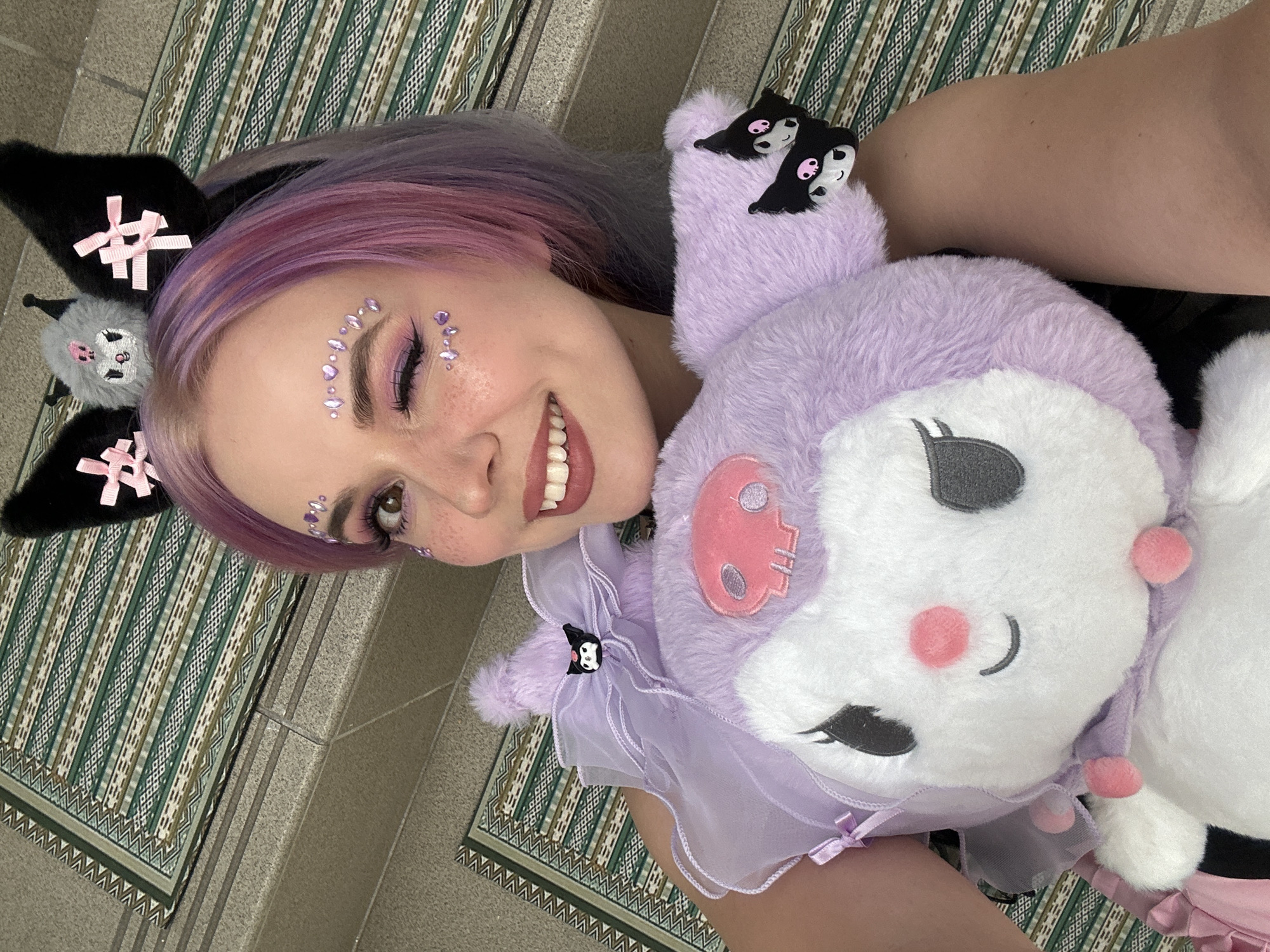 A young woman poses for a selfie. She has lavender hair, purple eye makeup, and rhinestones on her forehead and cheek. She is wearing black cat ear headbands with pink bows. The woman is holding a large plush toy, resembling Kuromi, a Sanrio character. The toy is lavender and white, with a tulle collar. The background shows a striped rug on a tiled surface with steps. The location is the city of Zagreb, Croatia.