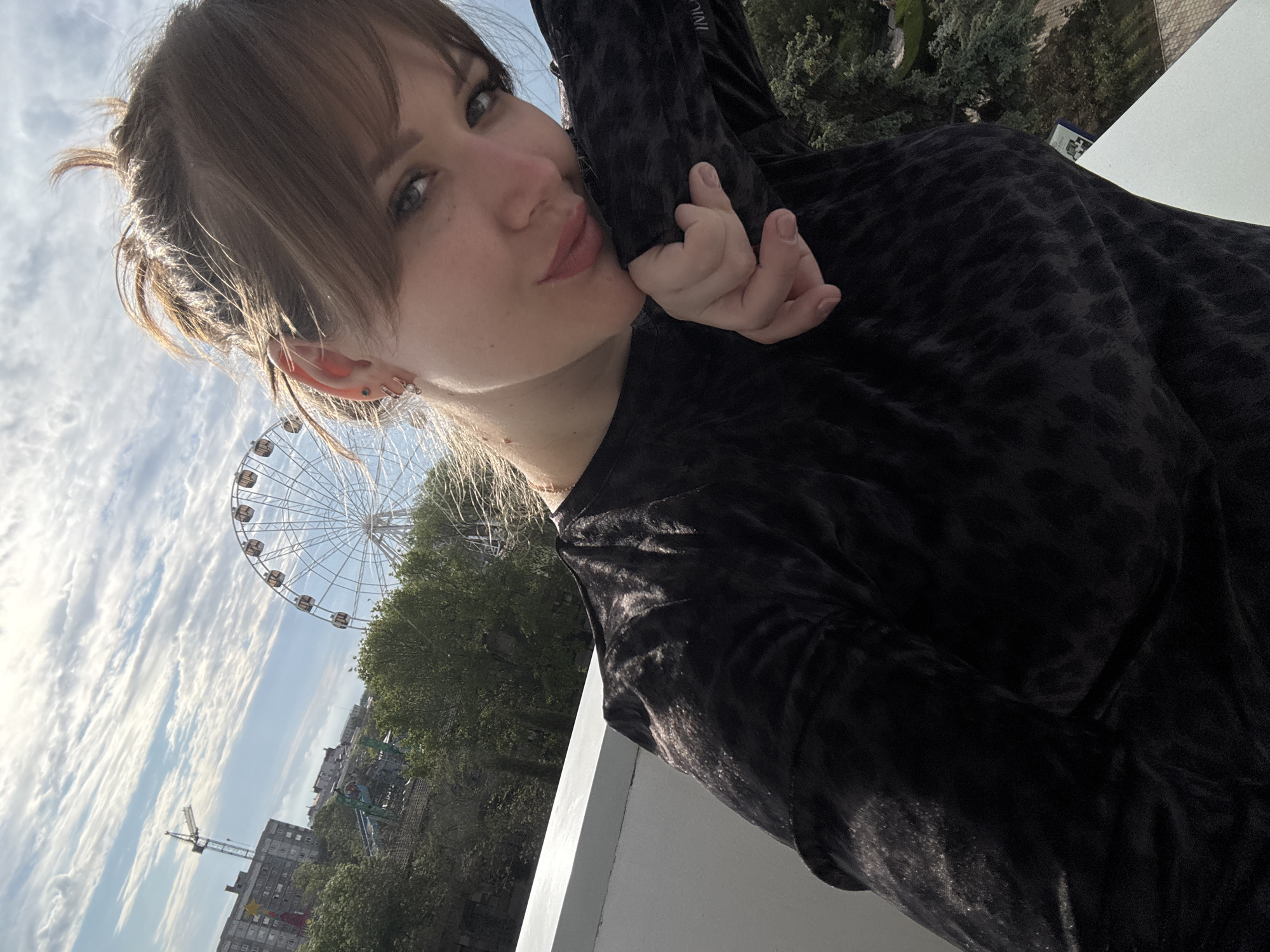 A medium shot captures a woman in a black dress with a subtle animal print, posing on a balcony. Her right hand is delicately touching her face. A Ferris wheel is visible in the background. The cityscape suggests Yevpatoriya, Ukraine, given the architectural style and the presence of the Ferris wheel as a landmark. The overall scene is outdoors during daylight, with trees, buildings, and construction cranes visible in the distant background.