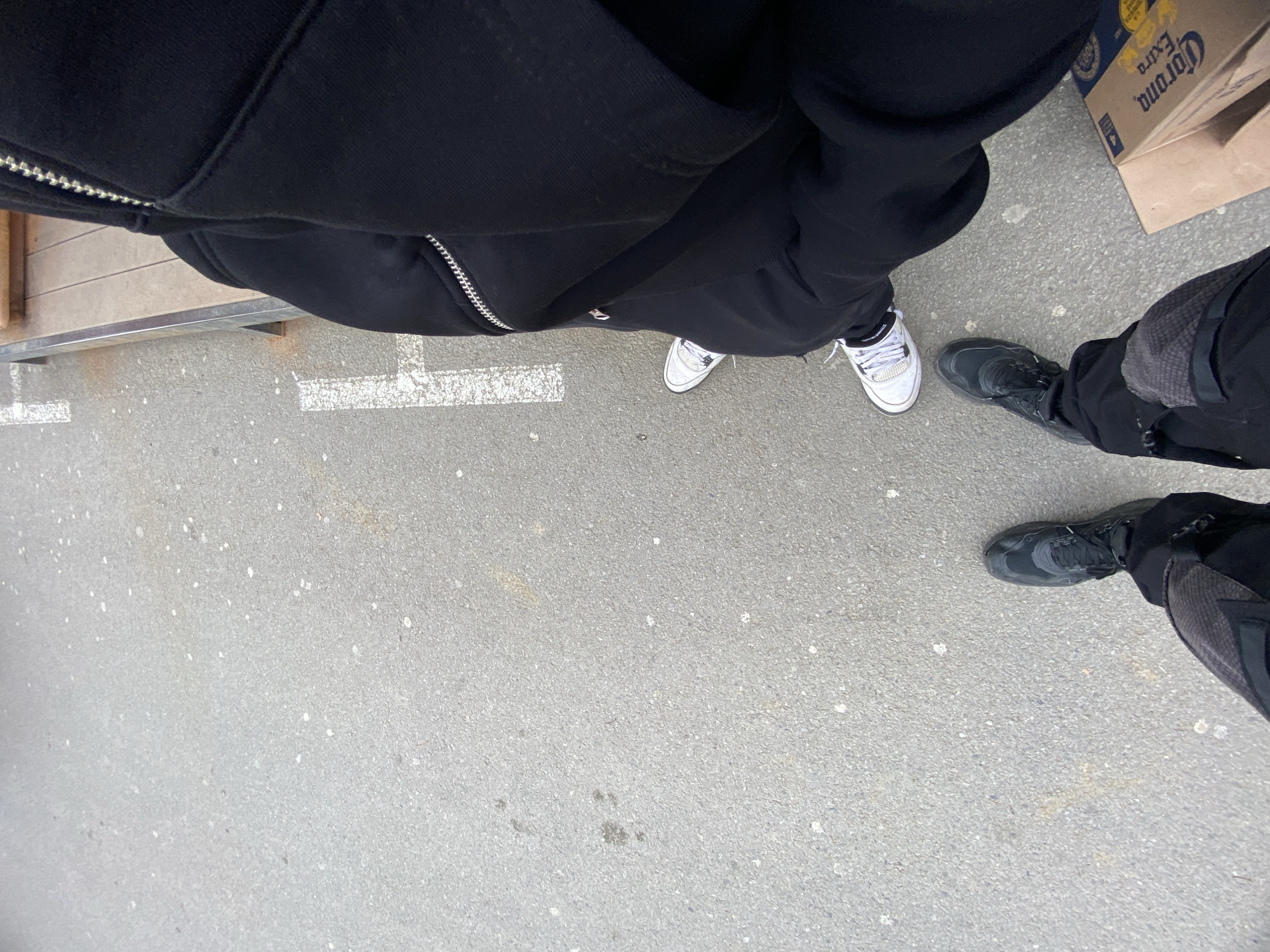 The image displays an overhead perspective focusing on the lower extremities of two individuals standing on a light grey asphalt surface in the city of Troyan, Bulgaria.
The individual on the right wears a black hooded sweatshirt with a prominent silver zipper down the front, black trousers, and white sneakers with black accents and white laces. Their stance is beside a wooden bench with a metallic frame, partially visible along the right edge of the frame. White painted lines, consistent with ground markings, are present on the asphalt near this individual.
The individual on the left wears black trousers, featuring what appear to be reinforced or textured panels on the thighs/knees, and dark grey/black athletic shoes with laces.
The asphalt ground shows minor surface irregularities, scattered white specks (consistent with small debris or dried residue), and a few darker smudges. In the bottom right corner, a partially collapsed cardboard box with "Corona Extra" branding is visible. Both individuals appear stationary.