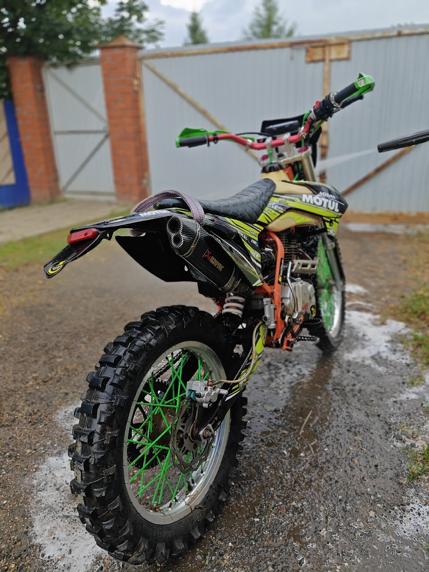 The image shows a dirt bike with green accents and a yellow gas tank parked on a wet, gravelly surface. The bike has a license plate that appears to be from Russia. In the background, there is a metal fence and two brick pillars, suggesting an outdoor setting, possibly a yard or driveway. The sky is cloudy. The bike is detailed with a "Motul" logo on the tank and an "Akrapovič" exhaust. The tires are knobby and appear to be wet. The overall scene suggests that the bike may have been recently washed or is being ridden in wet conditions. The location in Rassol'naya, Russia is noted.