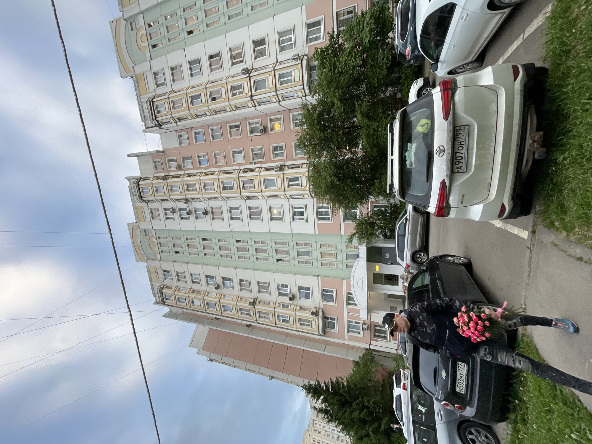The image shows a man carrying a bouquet of pink flowers walking on a sidewalk next to a row of parked cars in Moscow, Russia. The cars include a white Toyota SUV with license plate A907OH799 and a black car with license plate H503MY777. A tall apartment building is visible in the background. The sky is cloudy.