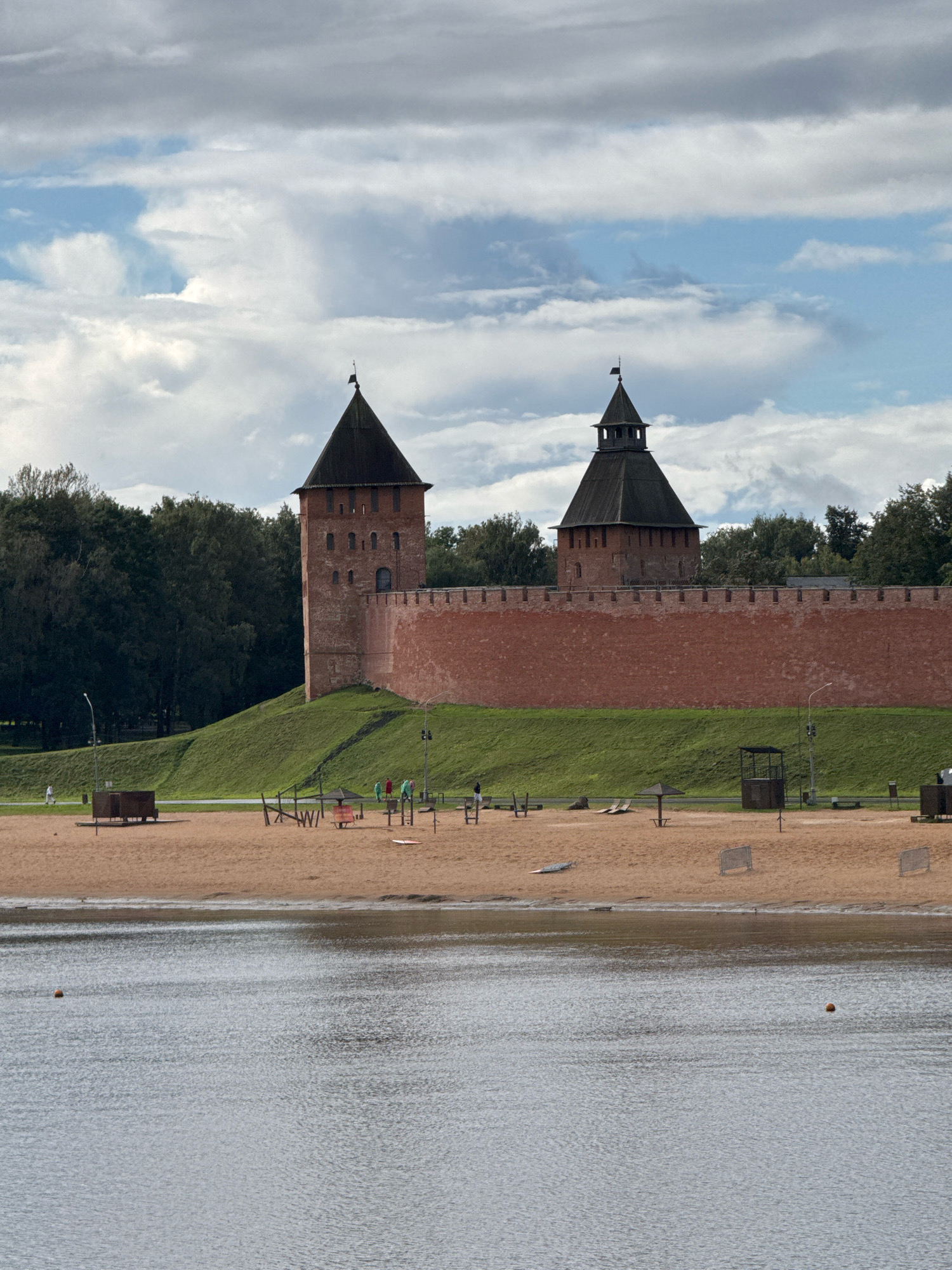 The image depicts a view of the Novgorod Kremlin in Veliky Novgorod, Russia, on a cloudy day. The Kremlin's red brick walls and two prominent towers dominate the background. In the foreground, there is a sandy beach area with various amenities, including beach chairs, umbrellas, and playground equipment. Several people are visible on the beach, some walking and others appearing to use the recreational facilities. The beach is situated alongside a body of water, likely a river, with ripples on its surface. Trees line the edge of the Kremlin grounds.
