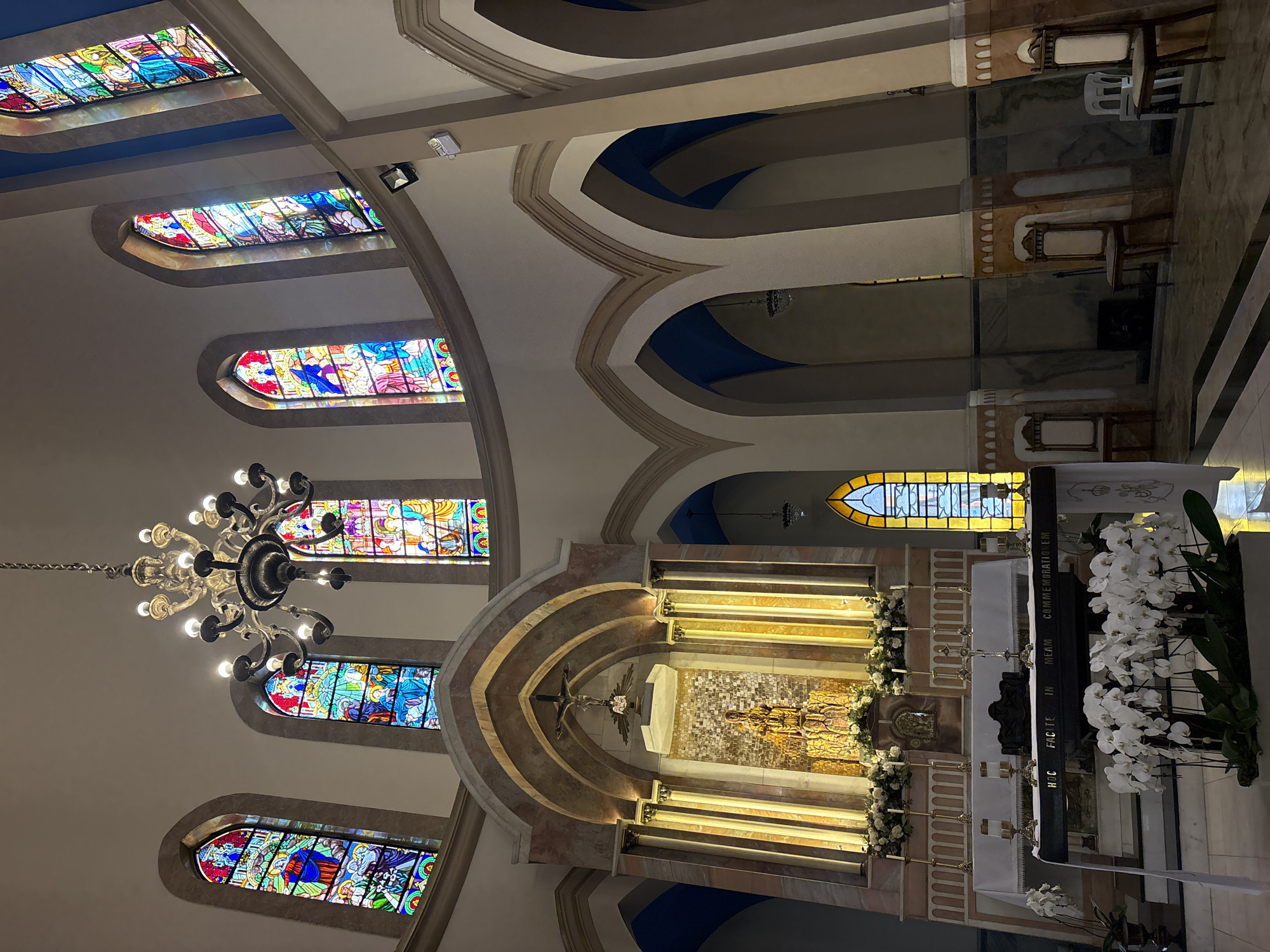 The image depicts the interior of the Igreja Nossa Senhora do Brasil, located in the city of São Paulo, Brazil. The church features a grand architectural design with soaring arched ceilings and tall stained-glass windows that illuminate the space with colorful light. At the front of the church is a beautifully adorned altar, featuring a mosaic backdrop, a statue of a religious figure, and a crucifix above. Flanking the altar are ornate columns and decorative lighting. The main body of the church is sparsely populated, with several rows of wooden chairs visible in the nave. A large chandelier hangs from the ceiling, adding to the ambiance. In the foreground, a prominent display of white orchids adds a touch of natural beauty to the solemn setting. The overall impression is one of reverence and architectural splendor.