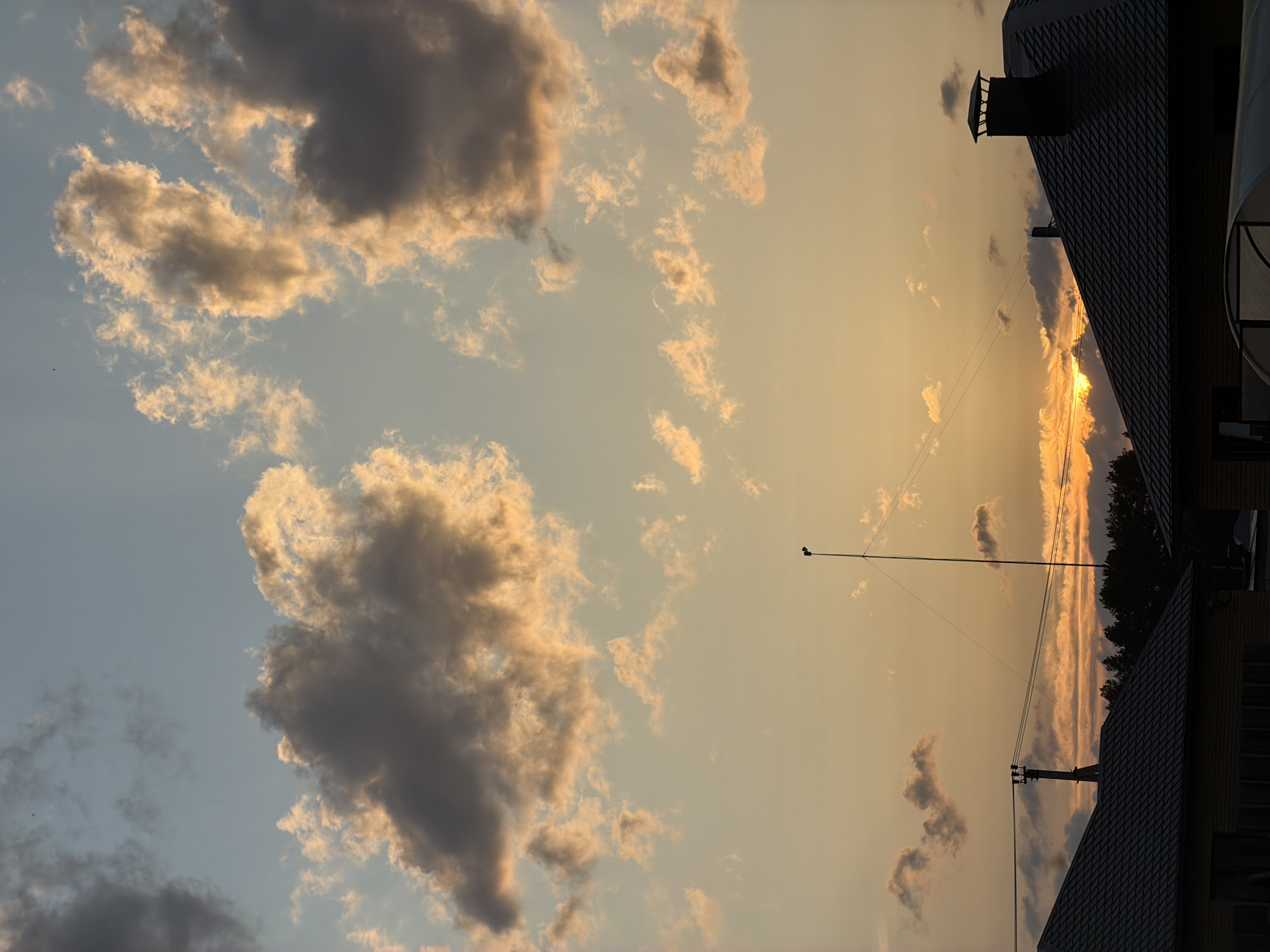 The image depicts a sky at sunset in Chaikovsky, Russia. The sky is a mix of pale blue and golden yellow, with scattered cumulus clouds. Some clouds are dark gray, while others are illuminated with a golden hue. A residential area is visible in the foreground. There is a rooftop and a chimney. In the middle ground, there are power lines, a utility pole, and a tall antenna. The sun is setting behind a line of trees on the horizon, creating a bright orange glow.