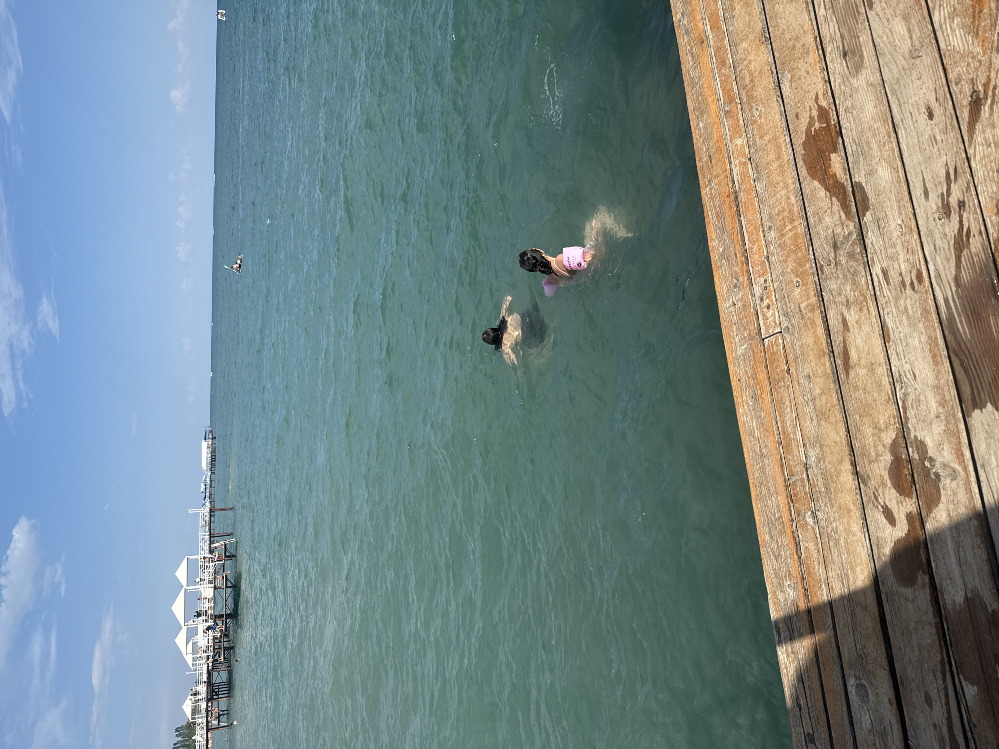 The image depicts a sunny day at a waterfront location, likely a beach or resort, in Sary-Oy, Kyrgyzstan. In the foreground, a wooden pier with weathered planks is visible, showing wet patches and shadows. The middle ground features two individuals swimming in the clear, greenish-blue water. One swimmer, wearing a pink flotation device, is positioned closer to the viewer, while another swimmer is further away. In the background, a white pier with white canopies extends into the water, and several people can be seen on it. Beyond the piers, the vast expanse of a body of water stretches to the horizon, meeting a bright blue sky dotted with white clouds. Trees line the distant shore. The overall scene conveys a sense of leisure and recreation on a warm day.