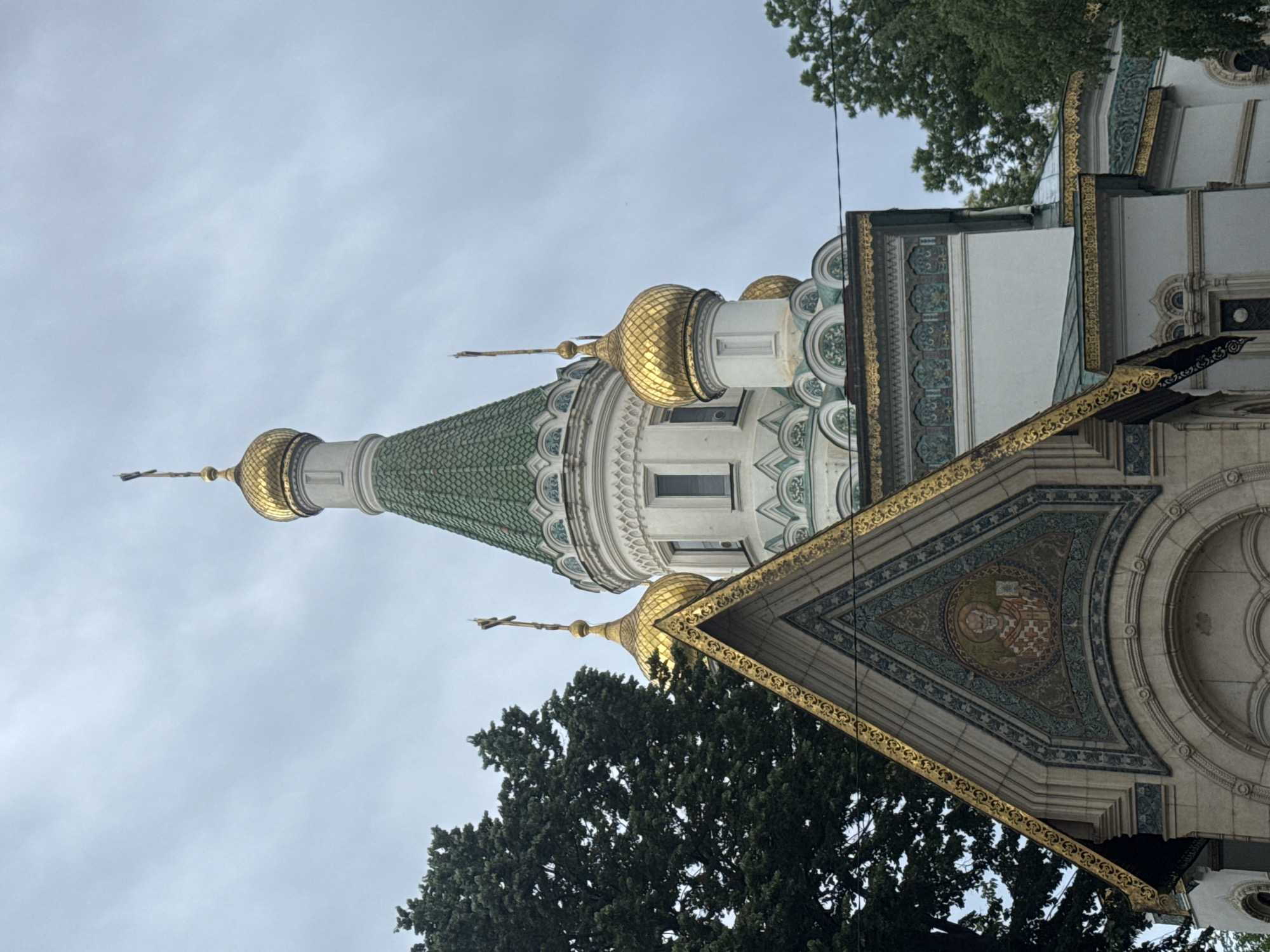 The image depicts a partial view of the Russian Church, officially known as the Church of St. Nicholas the Miracle-Worker, located in Sofia, Bulgaria. The architecture showcases a distinctive Russian Revival style. The scene includes portions of the church's exterior, highlighting the ornate decoration of a golden-domed roof with a green-tiled spire, and intricate mosaic work. The sky is overcast. Trees partially obscure the view.