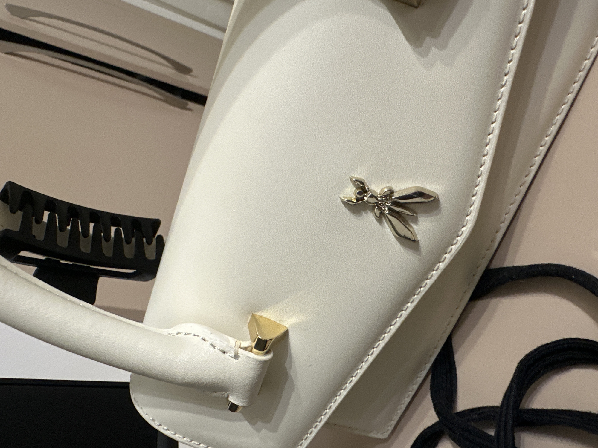 A close-up, high-angle shot captures a cream-colored handbag, likely made of textured leather or a similar synthetic material. The bag features a structured, curved top handle attached with gold-toned hardware, including a small, faceted pyramid-shaped stud on the left side. A distinctive gold-toned metal emblem, depicting a stylized figure with outstretched limbs (resembling a butterfly or dancer), is affixed to the front flap of the bag, positioned slightly below the horizontal midline. Visible contrast stitching outlines the edges of the flap and handle attachments. A black, coiled or braided strap is partially visible at the bottom left of the frame. The background consists of a light beige or tan surface, with a dark, serrated object (resembling a hair claw clip) partially visible behind the bag's handle. No people are present. The precise location of this scene in the city of Unknown, Unknown, cannot be determined from the image.