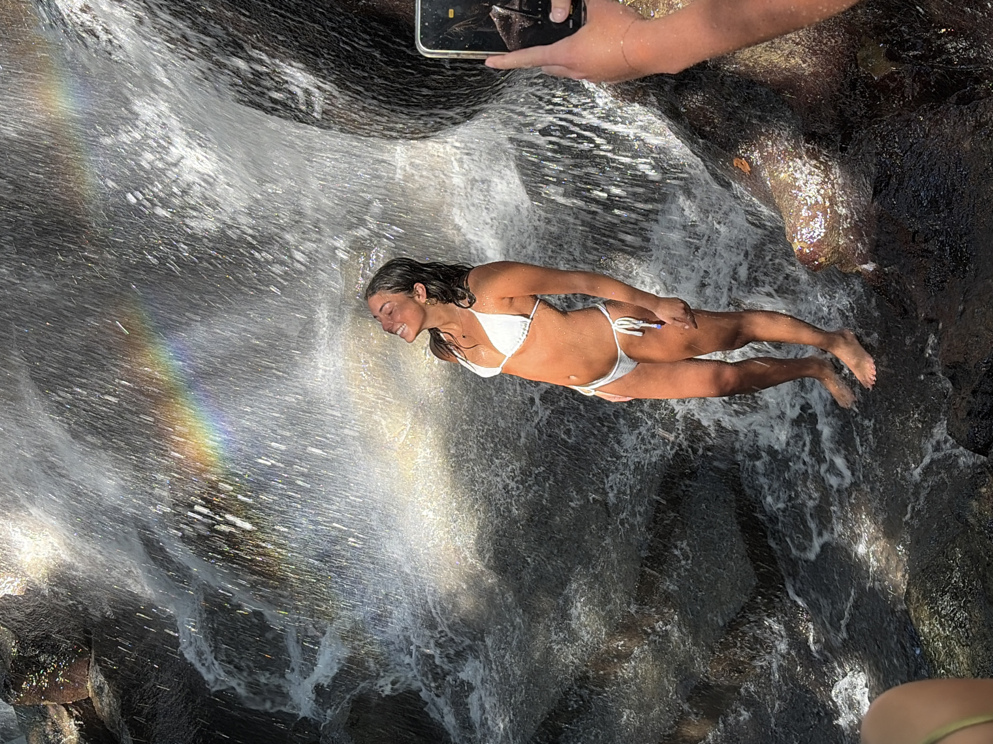 In the city of Gianyar, Indonesia, a young woman stands in a bikini under a waterfall, a rainbow visible in the mist. She is smiling with her eyes closed and her head tilted back. The water cascades down rocks, creating a dynamic and natural setting. Another person's hand is visible on the right side of the frame, holding a phone, suggesting a photo is being taken of the woman. The water is clear and abundant, indicating a natural water source like a waterfall or river. The surrounding rocks are dark and wet, typical of a waterfall environment.