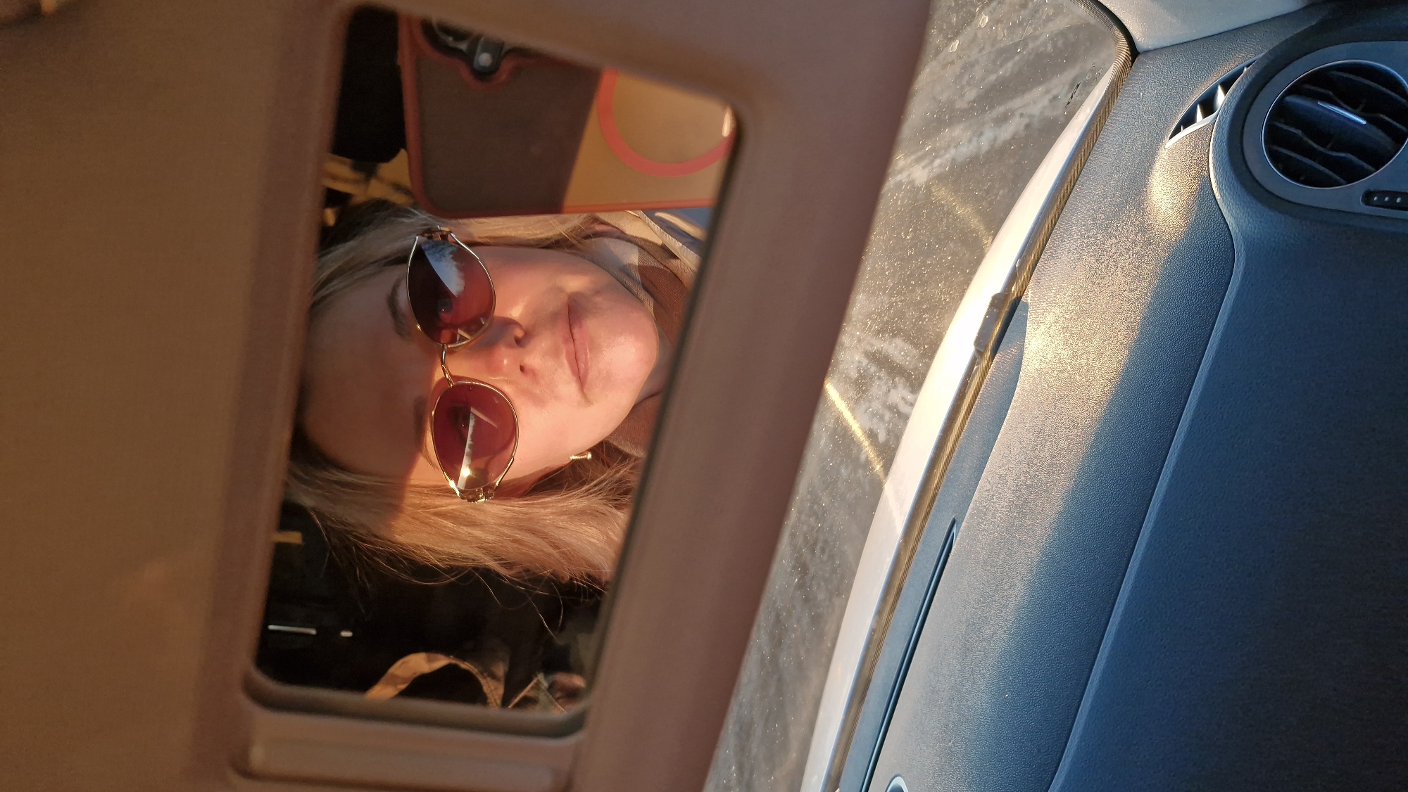 A woman with blonde hair and sunglasses is seen through a sunroof of a car. The interior of the car is visible, including the dashboard with an air vent. The image appears to have been taken at sunset or sunrise based on the lighting. The specific location in Otchor, Russia, is not discernible from the image.