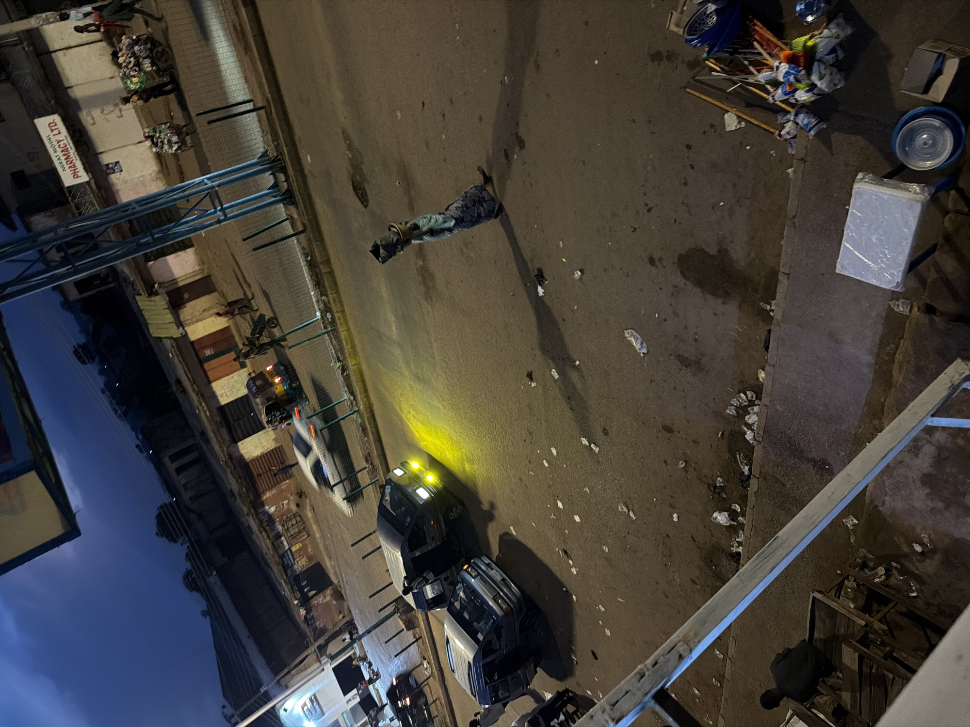 The image depicts a street scene at dusk in Bukuru, Nigeria. The sky is a deep blue, indicating the transition from day to night. Several vehicles are present, including a white car with its headlights on, a dark-colored van with yellow headlights illuminating the road, and another dark-colored van parked on the left side. A woman is walking in the middle of the street, carrying a basket on her head. There are buildings lining the street, some of which appear to be shops, including a sign that reads "NEPAL MODEL PHARMACY LTD." A few people are visible in the background, some pushing carts loaded with goods. Debris, such as plastic bags and papers, is scattered on the ground. The street is paved, and a sidewalk is visible on the right side of the image with cleaning supplies like brooms and mops. The overall impression is of a busy, active street in an urban environment during the evening.