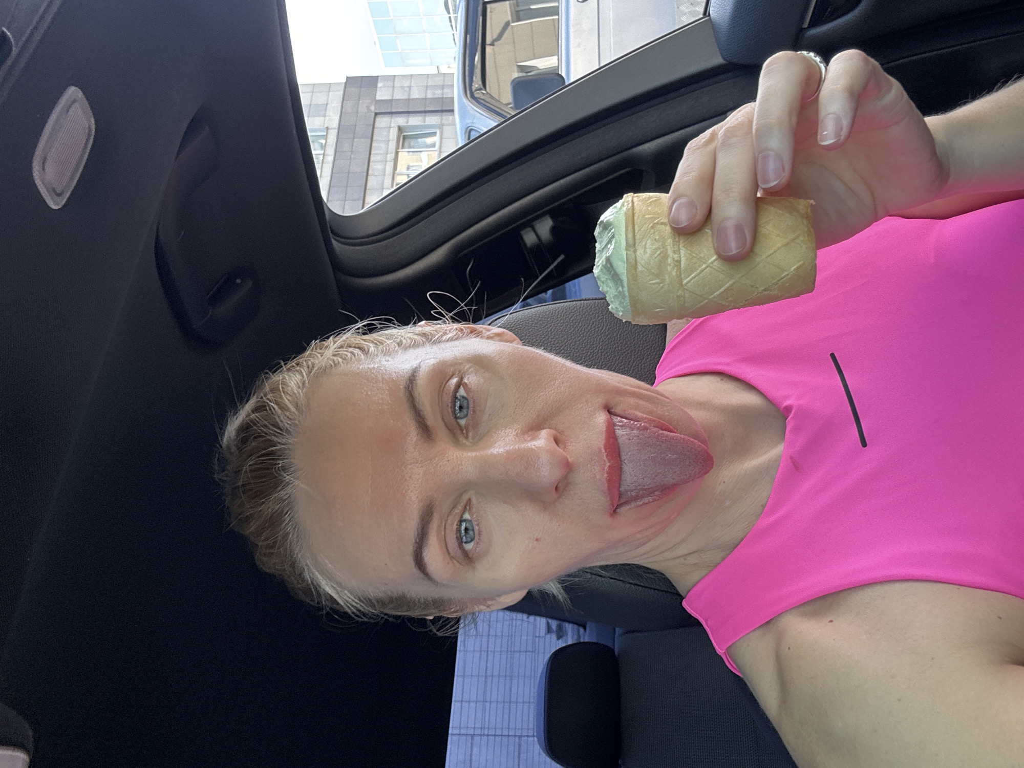 A blonde woman with blue eyes is sitting in the driver's seat of a car, sticking her tongue out towards the camera. She is wearing a bright pink top. In her right hand, she holds a wrapped ice cream cone. The car's interior is black. Through the car's window, buildings can be seen. The location is Unknown, Unknown.