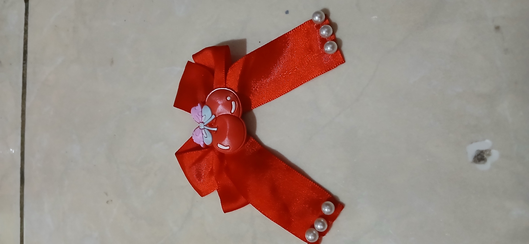 A close-up, high-angle shot shows a red satin bow adorned with a cherry charm and small pearl-like beads. The bow is centered on a light-colored tiled surface. The tiles have subtle variations in color and texture, with visible grout lines. There are a few dark specks and a small, indistinct mark on the tiles, suggesting a mundane environment. The image is static and does not depict any people or actions. The lighting is even, with no harsh shadows.