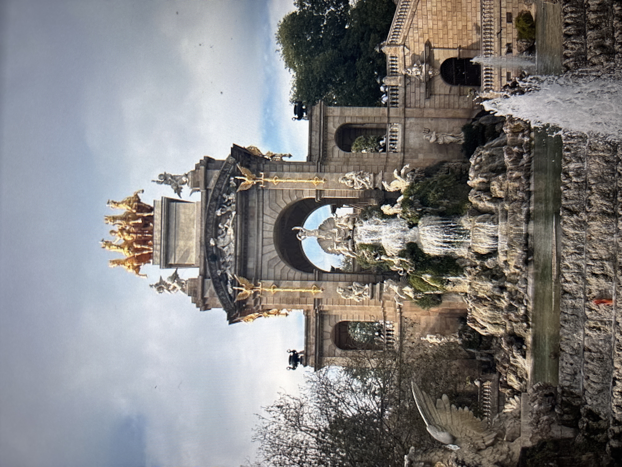 The image shows the monumental fountain of Parc de la Ciutadella in Barcelona, Spain. The fountain features a cascading waterfall, intricate sculptures of mythological figures, and is topped by a gilded chariot pulled by horses. The architecture is ornate, with arched doorways and classical columns. The scene is set against a partly cloudy sky and surrounded by trees. There are no people visible in the image. The description about Sofia, Bulgaria is incorrect; this is a landmark in Barcelona, Spain.
