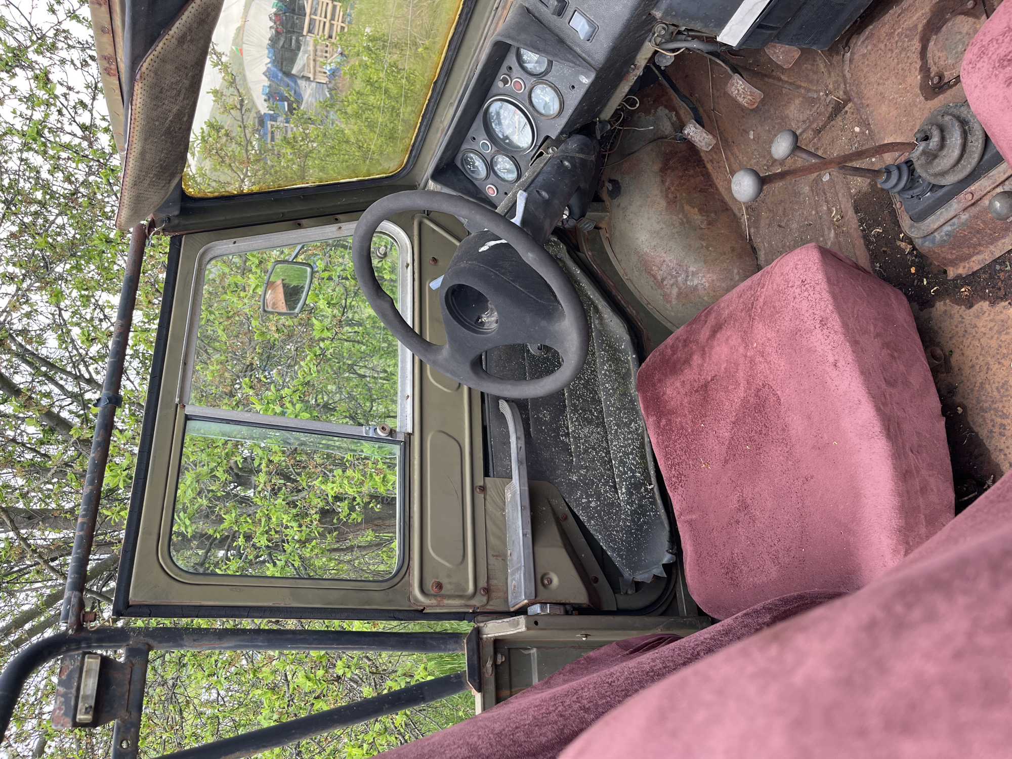 The interior of a heavily rusted, olive-green vehicle, likely a truck or off-road vehicle, is shown. The steering wheel is black and cracked. The dashboard is filled with gauges and displays signs of wear. The floor is severely rusted. Two gear shift levers are visible. The driver's seat is upholstered in a faded red material. The door is open, revealing green foliage outside. The door frame is olive green. A black roll cage is visible outside the door. The location, Lebnitsa, Bulgaria, is not definitively discernible from the image alone.
