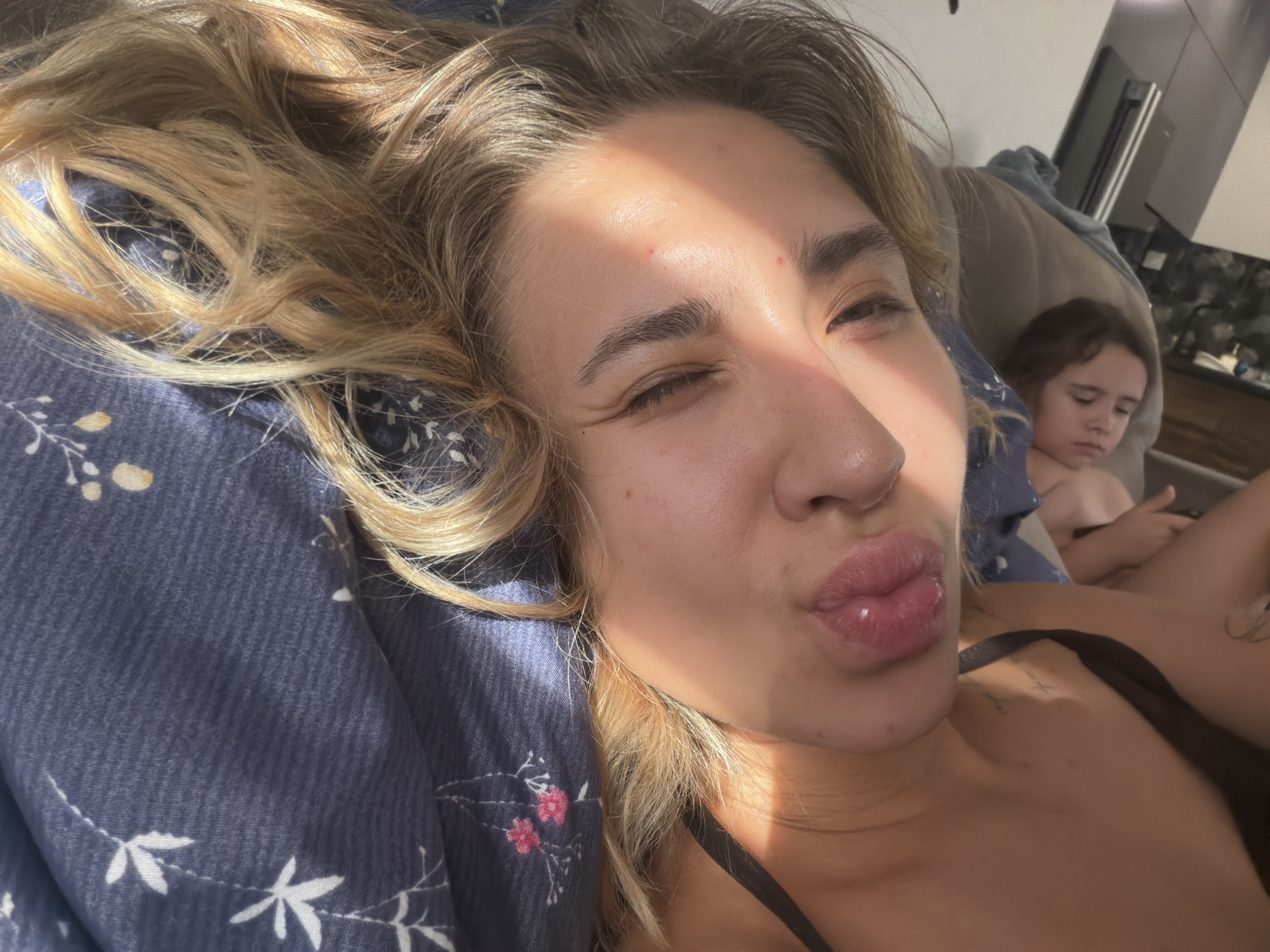 A close-up selfie shows a woman with blonde hair lying down, with her face turned towards the camera. She is winking with her left eye and puckering her lips. Sunlight is casting a warm glow across her face. In the background, a child is visible, lying down and looking away from the camera. The setting appears to be indoors, with a dark blue patterned pillow or blanket visible in the foreground. The scene is likely a personal moment, captured at home, possibly in Kamenka, Russia, though there is no specific visual information to confirm the location.