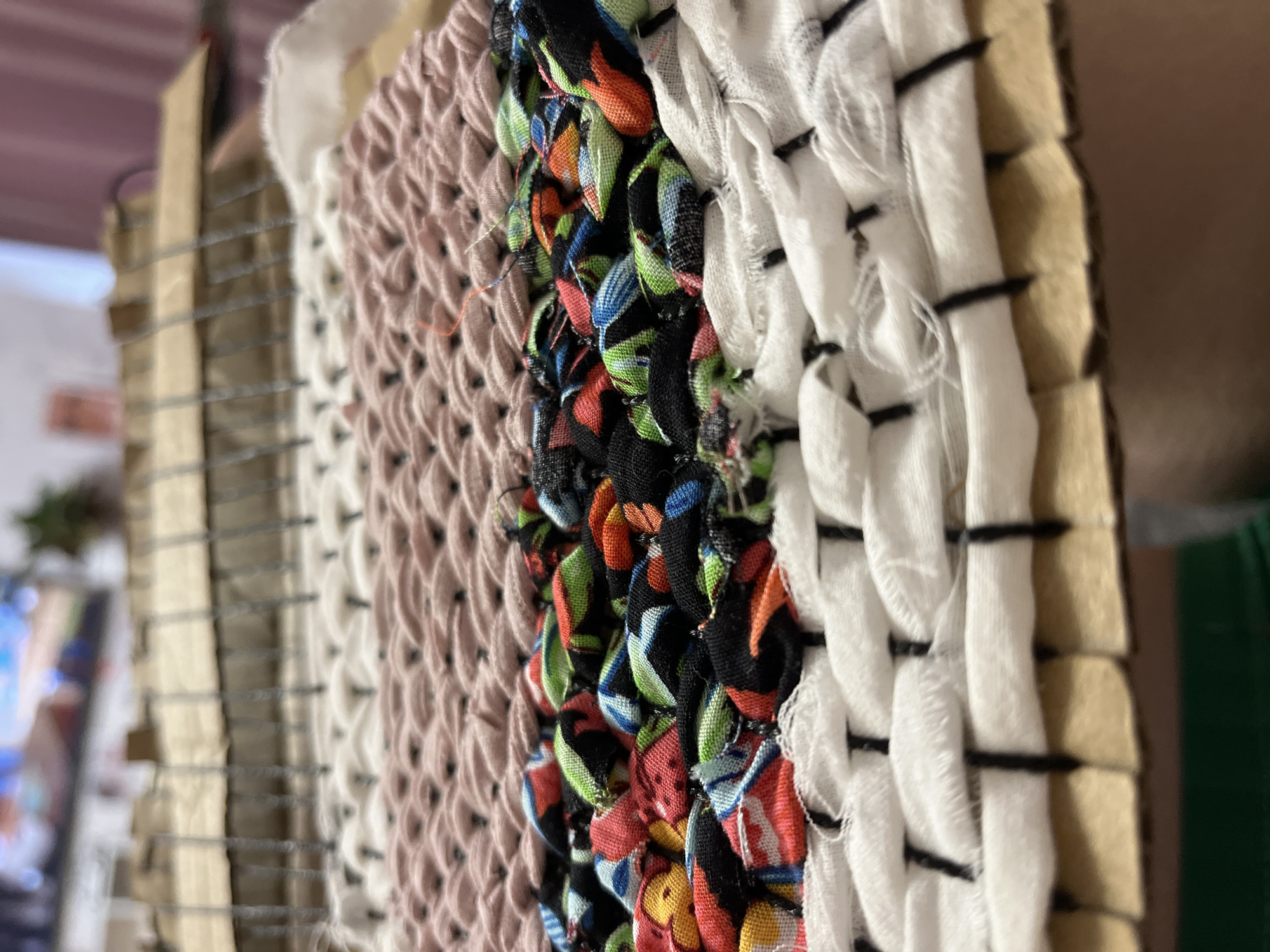 This image shows a close-up of a weaving project. The project features several horizontal bands of fabric, created using different techniques. The lowest band is made of white fabric woven with vertical black threads. Above this is a band of colorful fabric strips, appearing to be a patchwork or a brightly patterned material, woven in a looped or knotted fashion. Next is a band of solid pink fabric, also woven in a patterned manner, possibly a knit or crochet stitch. At the top is a band of white fabric with a looser weave, showing the warp threads underneath. The weaving is being done on a loom constructed from cardboard and what appear to be metal rods or dowels. The background is blurred but suggests an indoor setting with soft pink curtains and furniture. There are no people visible in the image. The location of Krasnyye Barrikady, Russia is not discernible from the visual information in the image.