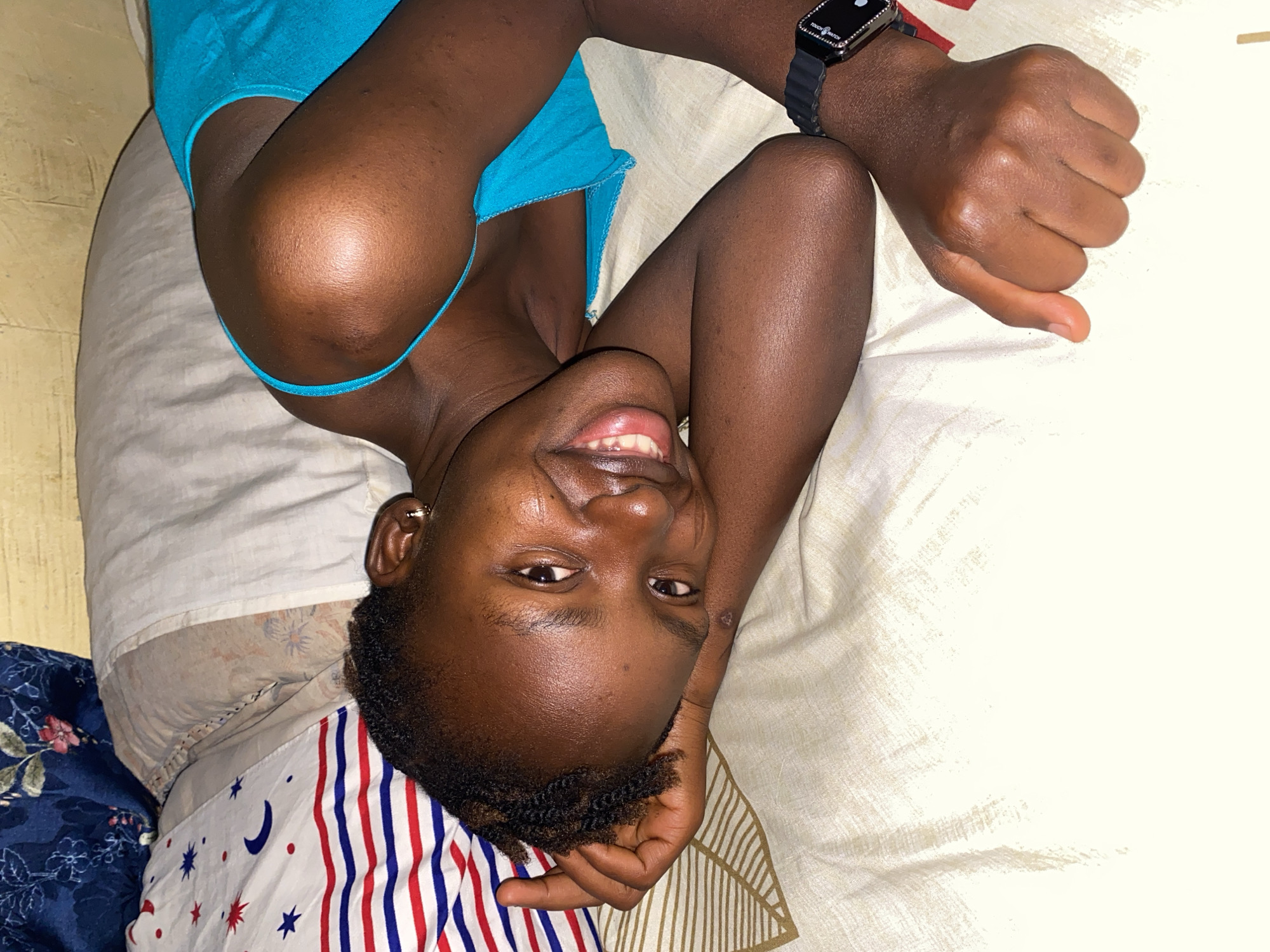 A dark-skinned woman is lying on a bed, smiling and looking at the camera. She wears a blue tank top and a black smartwatch. The bed has a white sheet with a patterned section on one side containing stars, moons, and red and blue stripes. The scene is indoors. The city of Kuru Dungus, Nigeria is not discernible from the image.