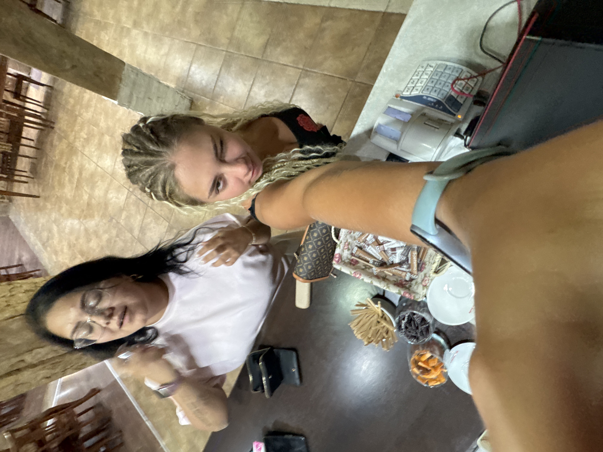 In a scene captured from a high-angle perspective, two women are present. The woman on the left, wearing glasses and a light pink t-shirt, appears to be speaking with her eyes closed and hands raised. The woman on the right, with blonde braided hair, winks at the camera while wearing a black tank top with a red floral design. They are situated behind a dark brown counter.
On the counter, several items are visible: a black wallet, a small brown patterned bag, a tray filled with individually wrapped sugar packets, a small white bowl containing small dark objects, another small white bowl with what appears to be dried fruit, a stack of thin brown sticks, and two white saucers. To the right of the counter, a calculator with a pink cord is present, along with a dark tablet device.
The background suggests an indoor setting with tiled flooring and wooden architectural elements, possibly indicative of a cafe or restaurant. The lighting is moderate, casting some shadows. The overall impression is a casual moment shared between two individuals. The image does not contain any explicit references to Sredets, Bulgaria.
