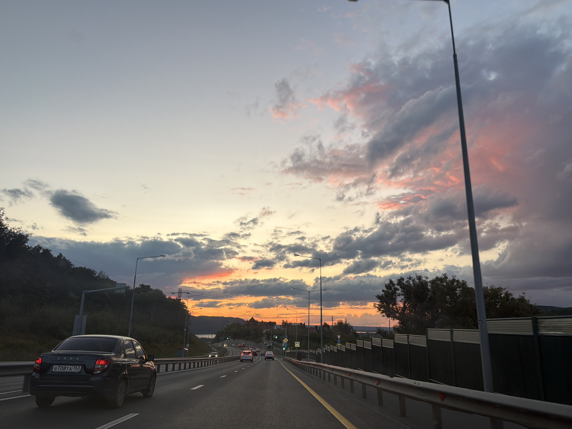 The image captures a view from a moving vehicle on a multi-lane highway in Samara, Russia, during sunset. The sky is a dramatic display of color, with clouds painted in hues of pink, orange, and gray against a fading blue. The sun is low on the horizon, casting a warm glow.
In the foreground, a dark-colored Lada sedan is driving in the right lane. Its license plate is visible, reading "B708TA 763". Several other vehicles are visible further down the highway, some with their headlights on. Streetlights and traffic signals are present along the roadside.
To the left of the highway, a forested hillside rises, its dark silhouette contrasting with the illuminated sky. A body of water, possibly the Volga River, is visible in the distance on the right, beyond a series of low buildings and trees. A metal guardrail runs along the side of the road, and a dark, corrugated fence is erected behind it. The road itself is paved and marked with white and yellow lines. The overall scene suggests a journey on the outskirts of Samara as day turns into night.