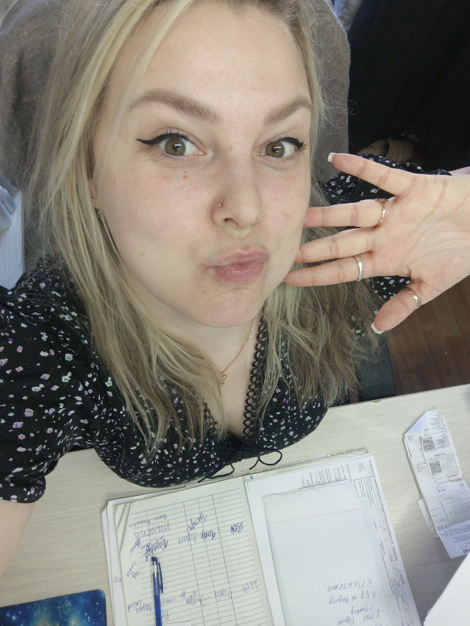A selfie of a fair-skinned blonde woman with green eyes and a nose piercing, wearing a black floral print top, at a desk. The woman is puckering her lips and holding up her hand which has multiple rings. There are papers, receipts, and a pen on the desk. One of the papers has handwritten notes in Russian. The text "Пермь" is visible in the notes. The image was taken in Perm, Russia.