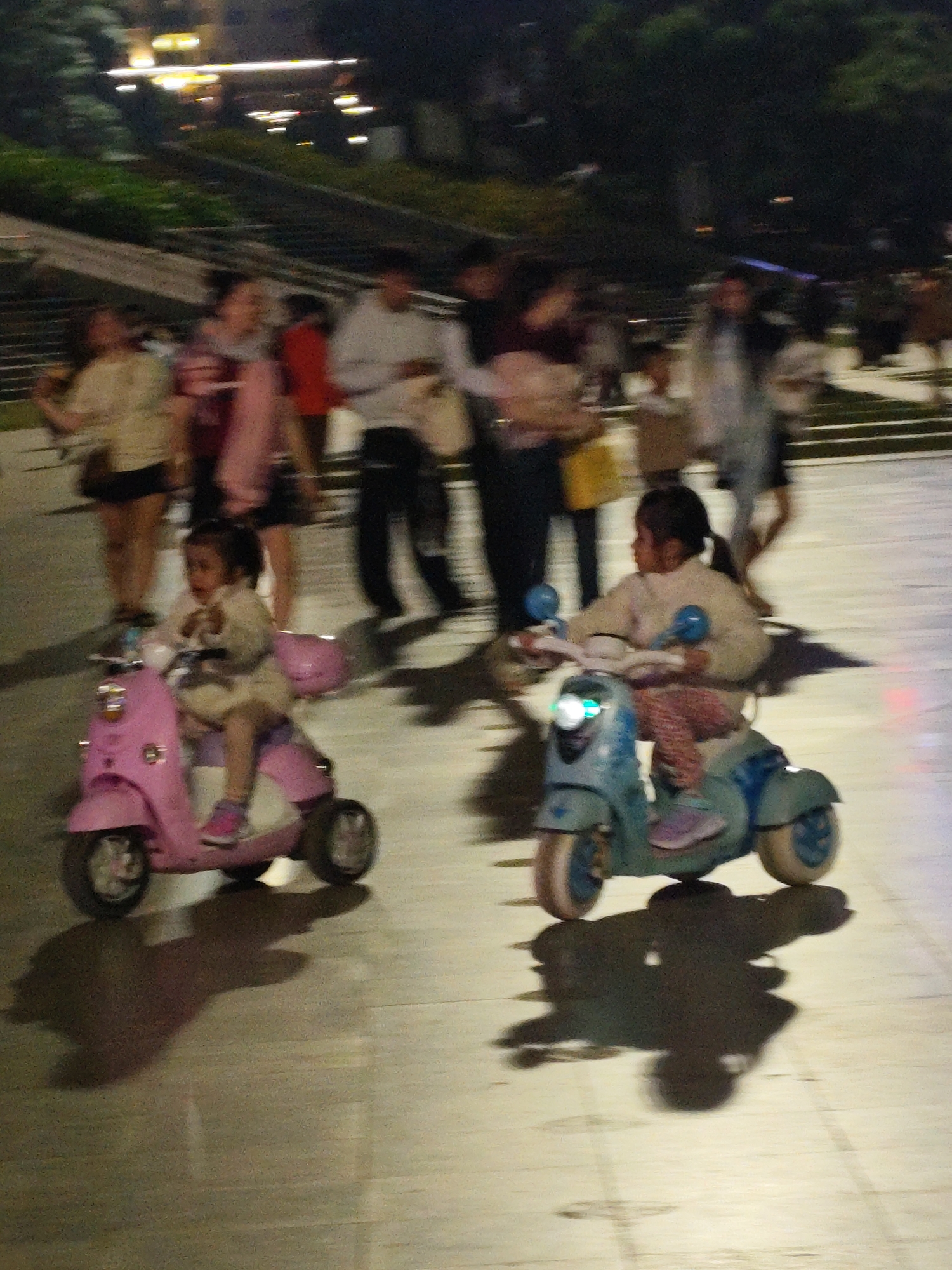 Two young girls are riding toy electric scooters on a paved outdoor area, likely a public plaza or walkway in Da Lat, Vietnam. The girl on the left is riding a pink scooter, and the girl on the right is riding a blue scooter. Both girls are wearing casual clothing and appear to be enjoying themselves. In the background, several adults are walking up a set of stairs. The scene is dimly lit, suggesting it is nighttime or late evening, and there are some artificial lights illuminating the area. The overall impression is one of family leisure and recreation in an urban setting.