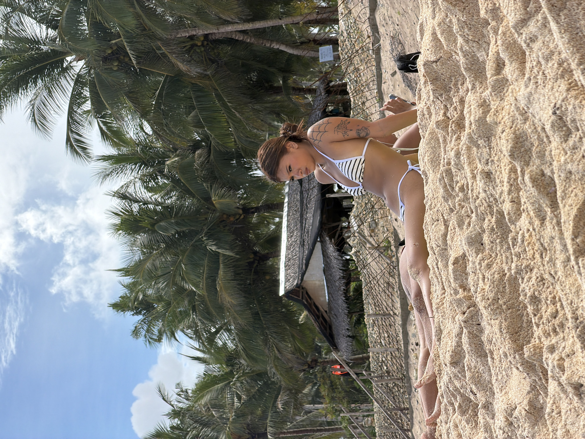 The image shows a person, possibly female, in a striped bikini standing on a sandy area, presumably a beach. The background reveals palm trees, clear skies with light clouds, and a small building constructed from natural materials, reminiscent of local Filipino architecture. The location is likely Pilar, Philippines, based on the description. The person appears to be holding a pen or thin object in their right hand. The sand is coarse and textured. A section of wire fencing is visible near the sandy area.