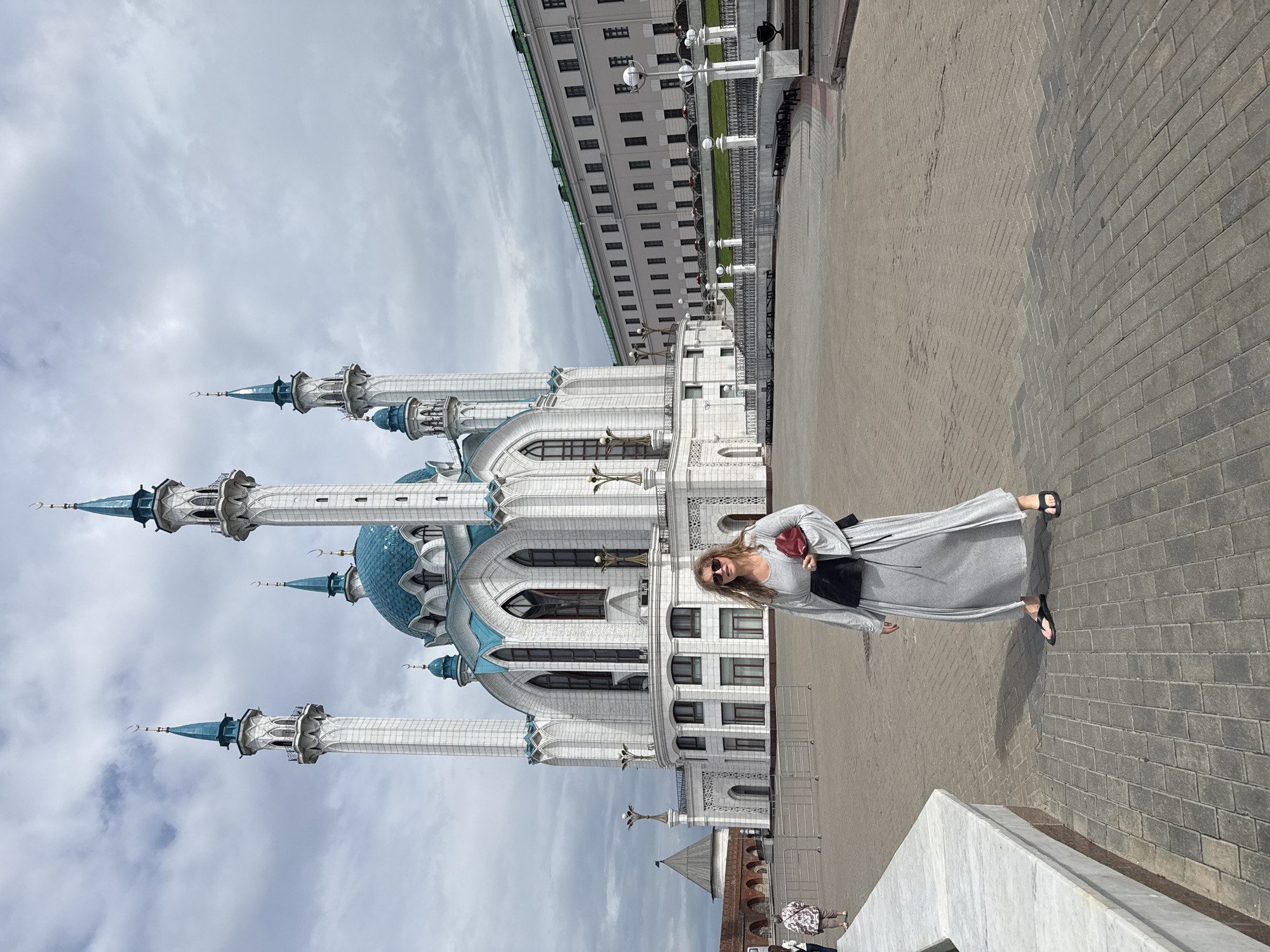 A woman in a gray maxi dress and black flip-flops stands in a paved plaza in front of the Kul Sharif Mosque in Kazan, Russia. The mosque, a white structure with a blue dome and multiple minarets, is illuminated by daylight. The woman holds a red clutch bag in her right hand. To the right of the mosque, a long, light-colored building with green trim stretches across the frame. The plaza is paved with interlocking gray bricks, and a low, white marble barrier is visible on the left side of the image. The sky is overcast with gray clouds.